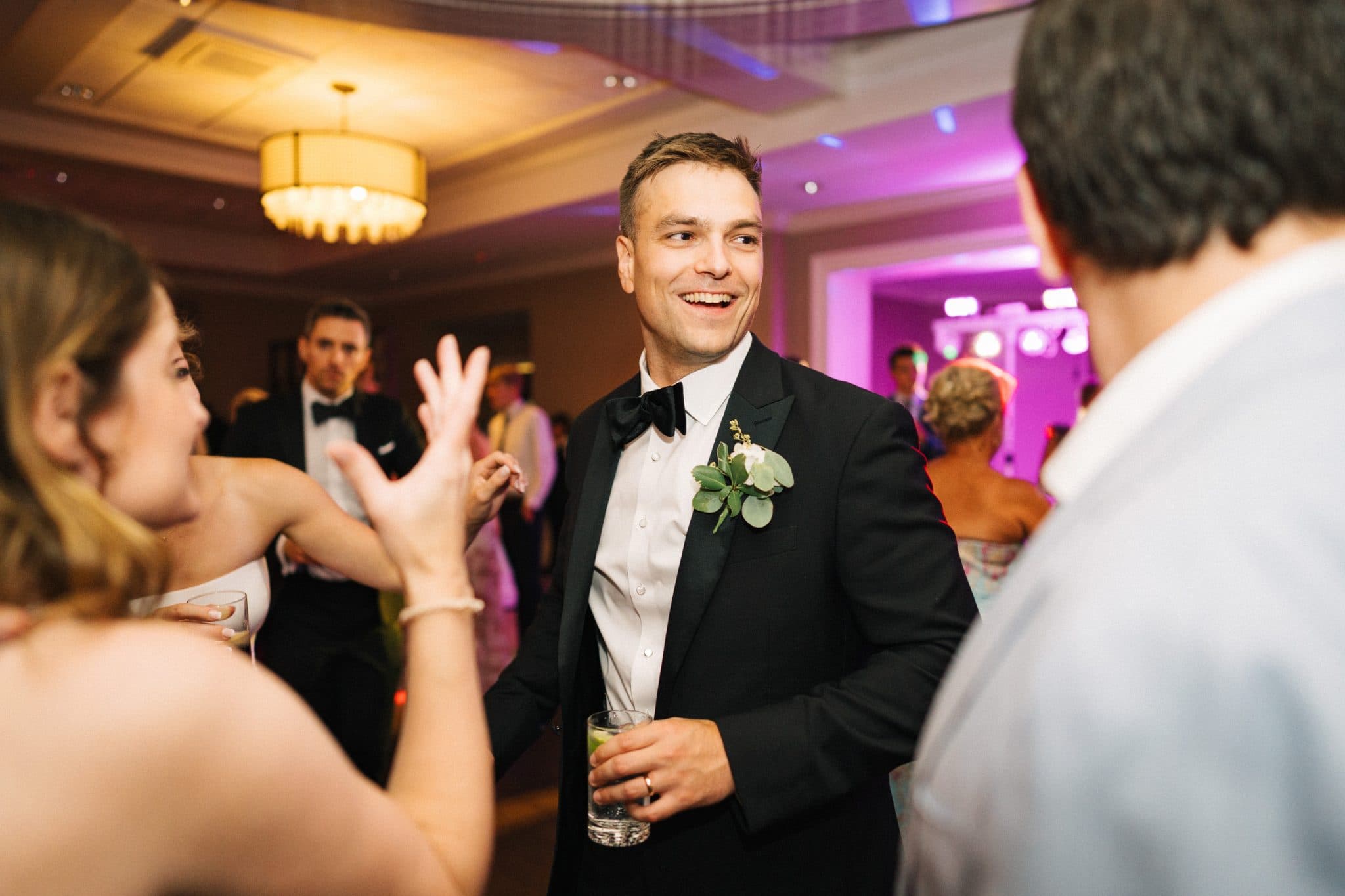 A man in a tuxedo with a boutonniere smiles while holding a drink, surrounded by guests at a wedding reception.