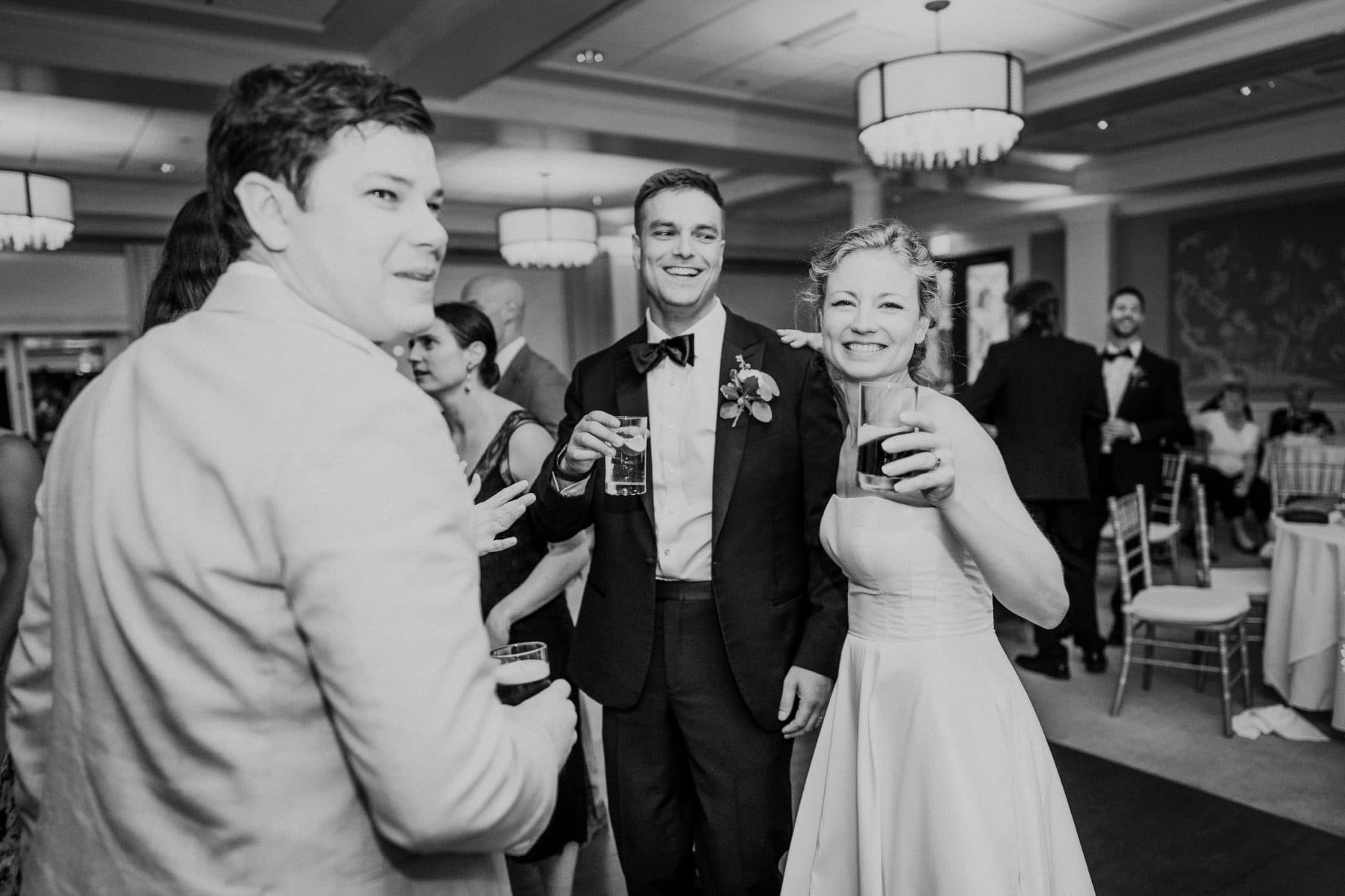 A bride and groom smile and hold drinks, surrounded by guests in a formal setting.