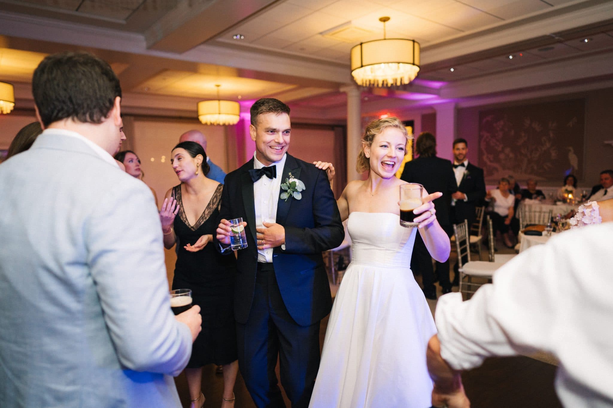A bride in a white dress and a groom in a tuxedo smile and hold drinks at a wedding reception.