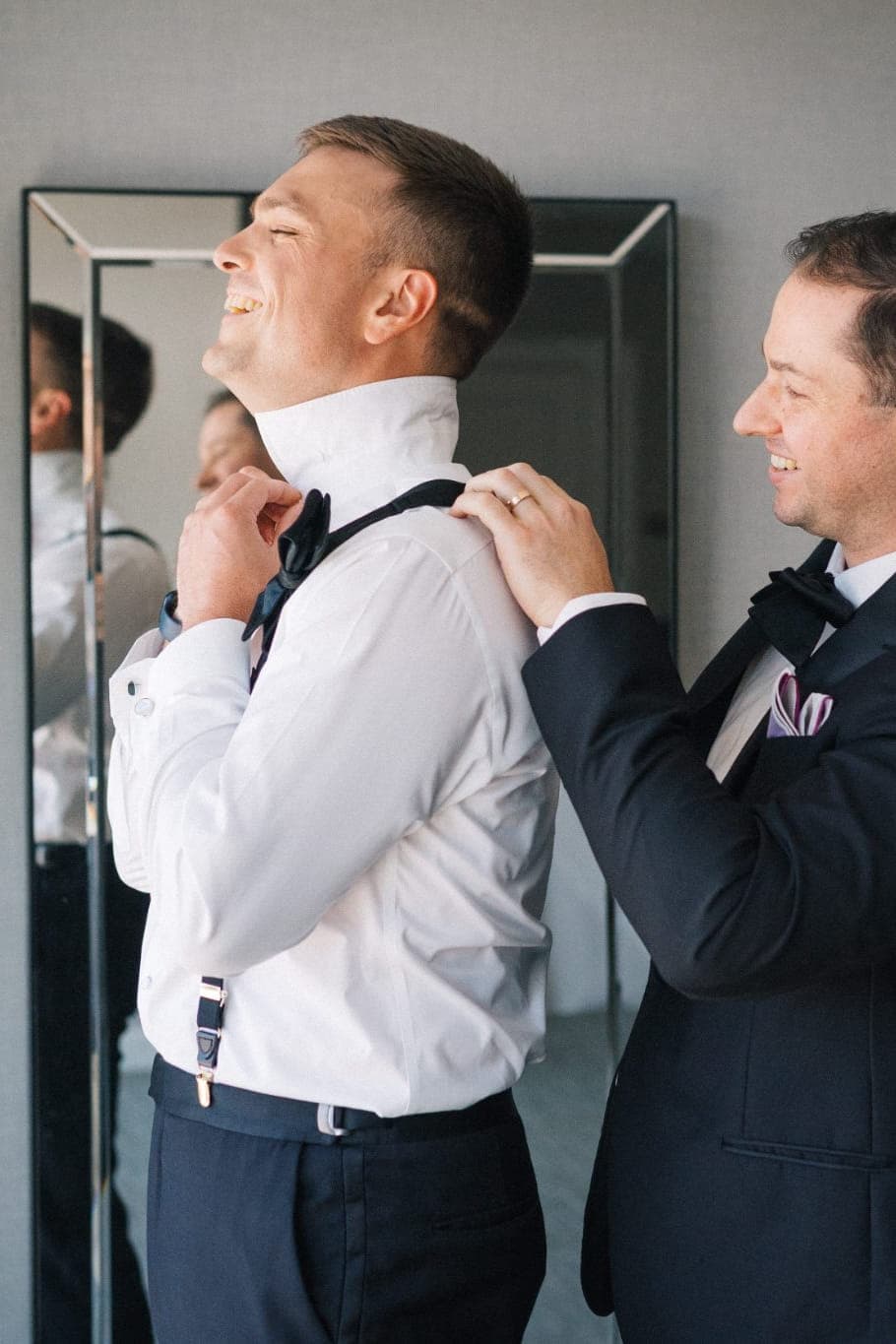 Two men adjust a white shirt and bow tie in front of a mirror.