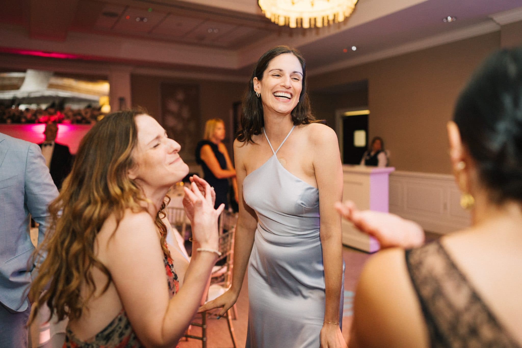 Two women in elegant dresses laugh and gesture at a lively indoor event.