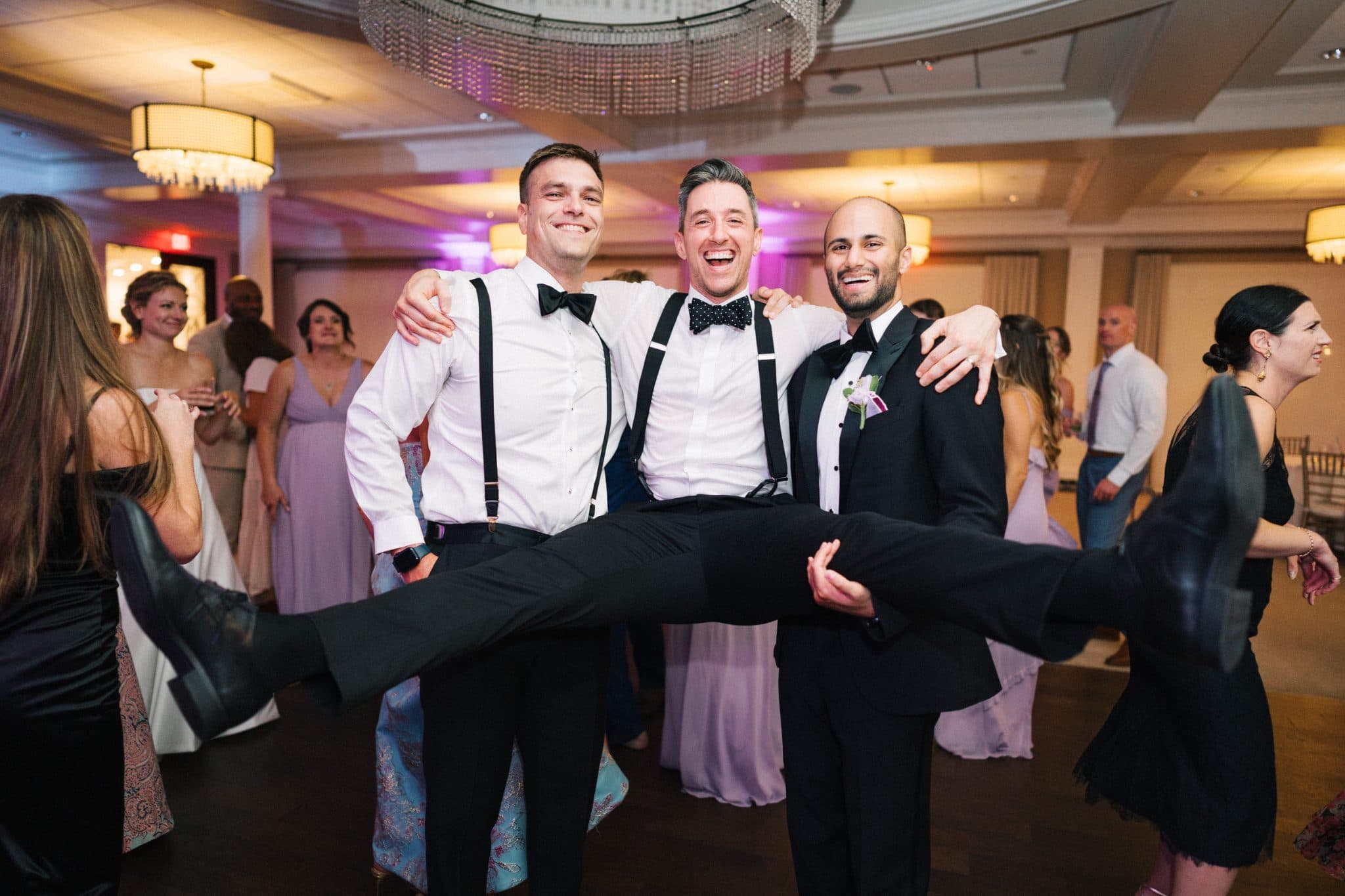 Three men in formal attire pose together with one man lifting another in a playful manner at a wedding reception.