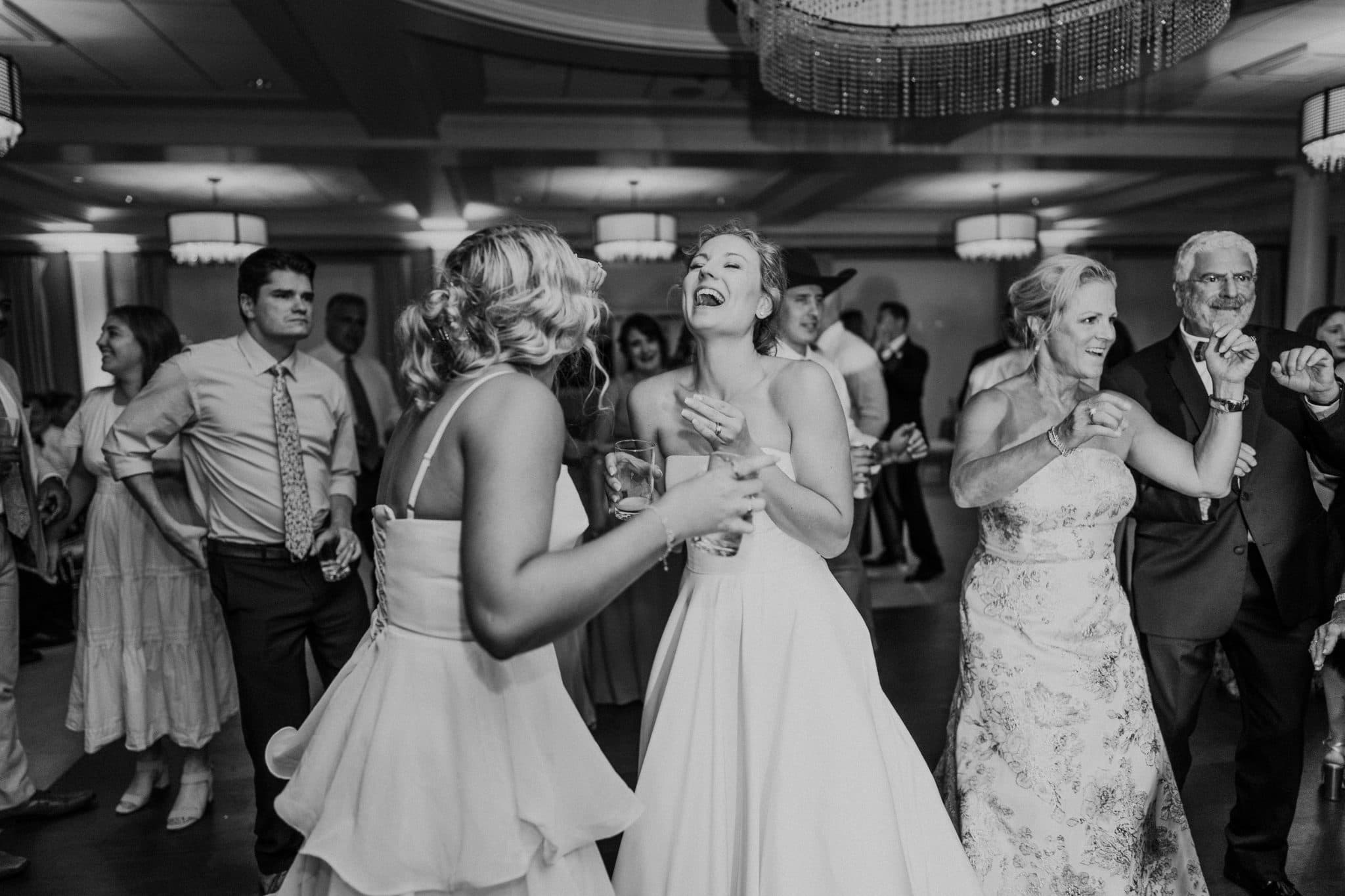 A bride dances joyfully with friends on a crowded dance floor, surrounded by guests in formal attire.
