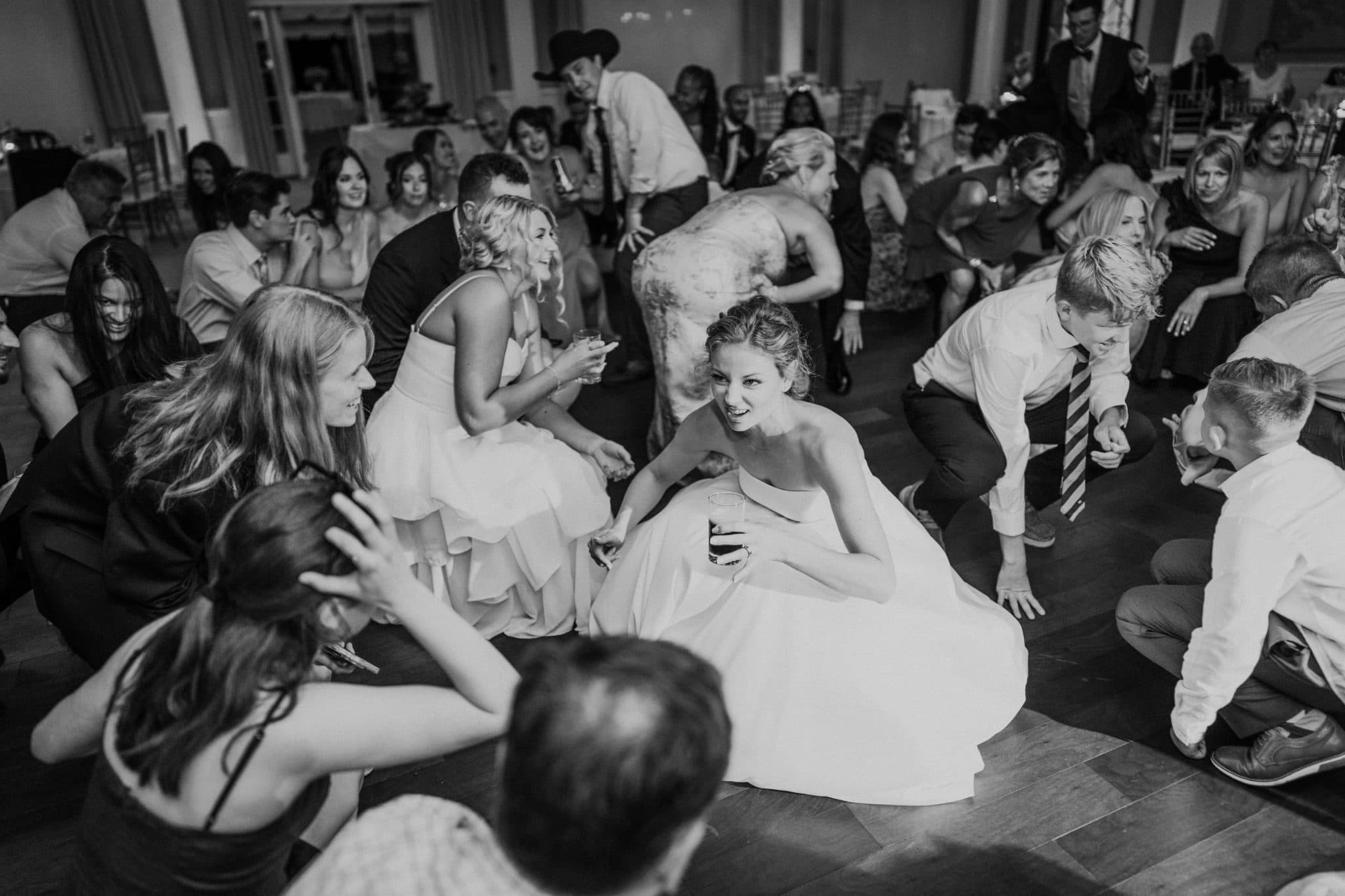 A bride sits on the dance floor surrounded by guests in a lively wedding reception.