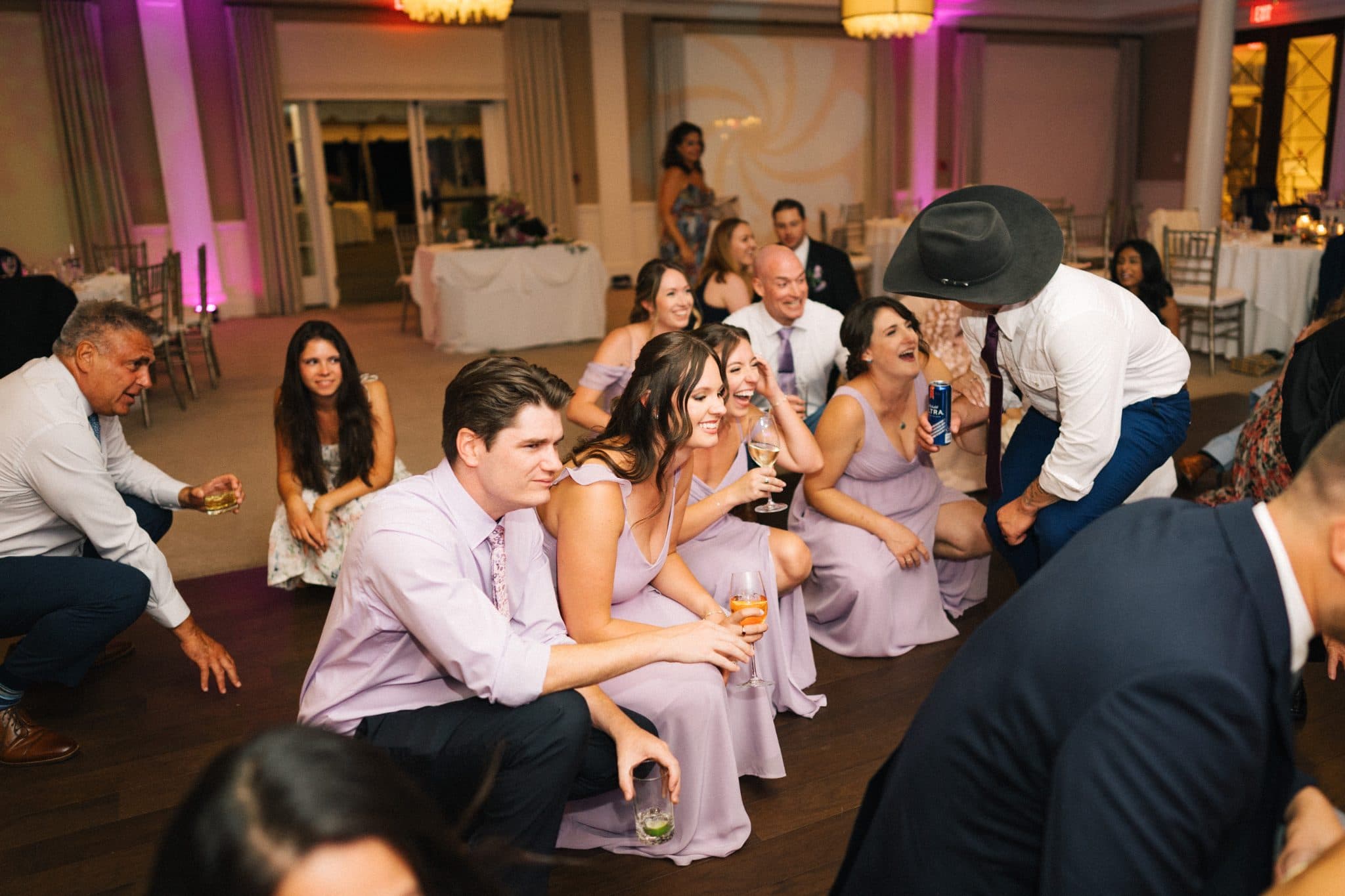 A group of people in a wedding setting, some seated and others standing, with drinks in hand, in a warmly lit room.