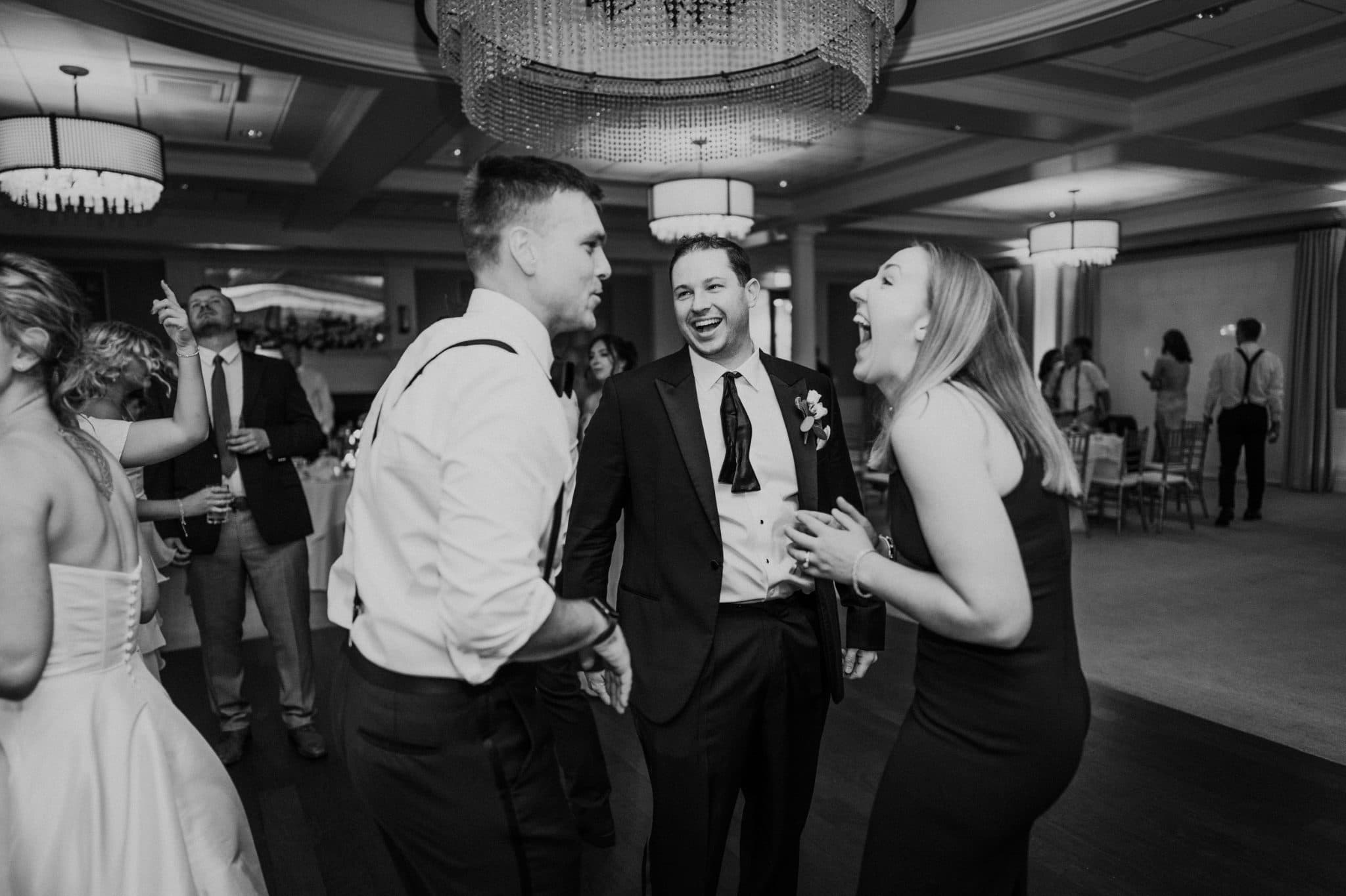 A black-and-white wedding scene shows three people in formal attire laughing and interacting in a grand hall with chandeliers.