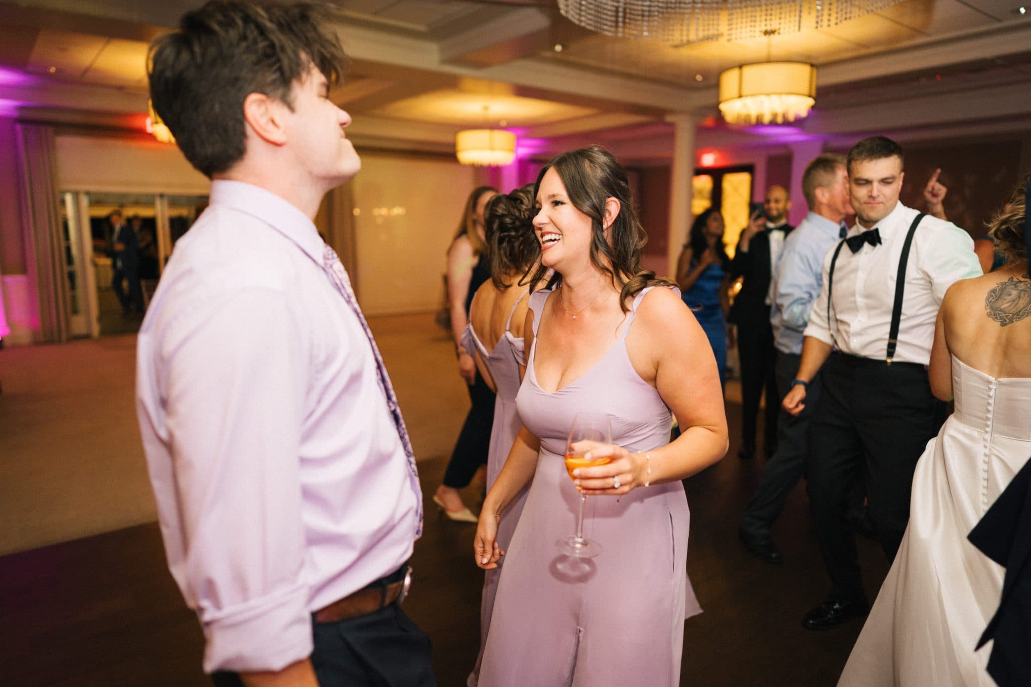 A woman in a lavender dress holds a glass of orange liquid, smiling at a man in a light purple shirt and tie at a lively indoor event.