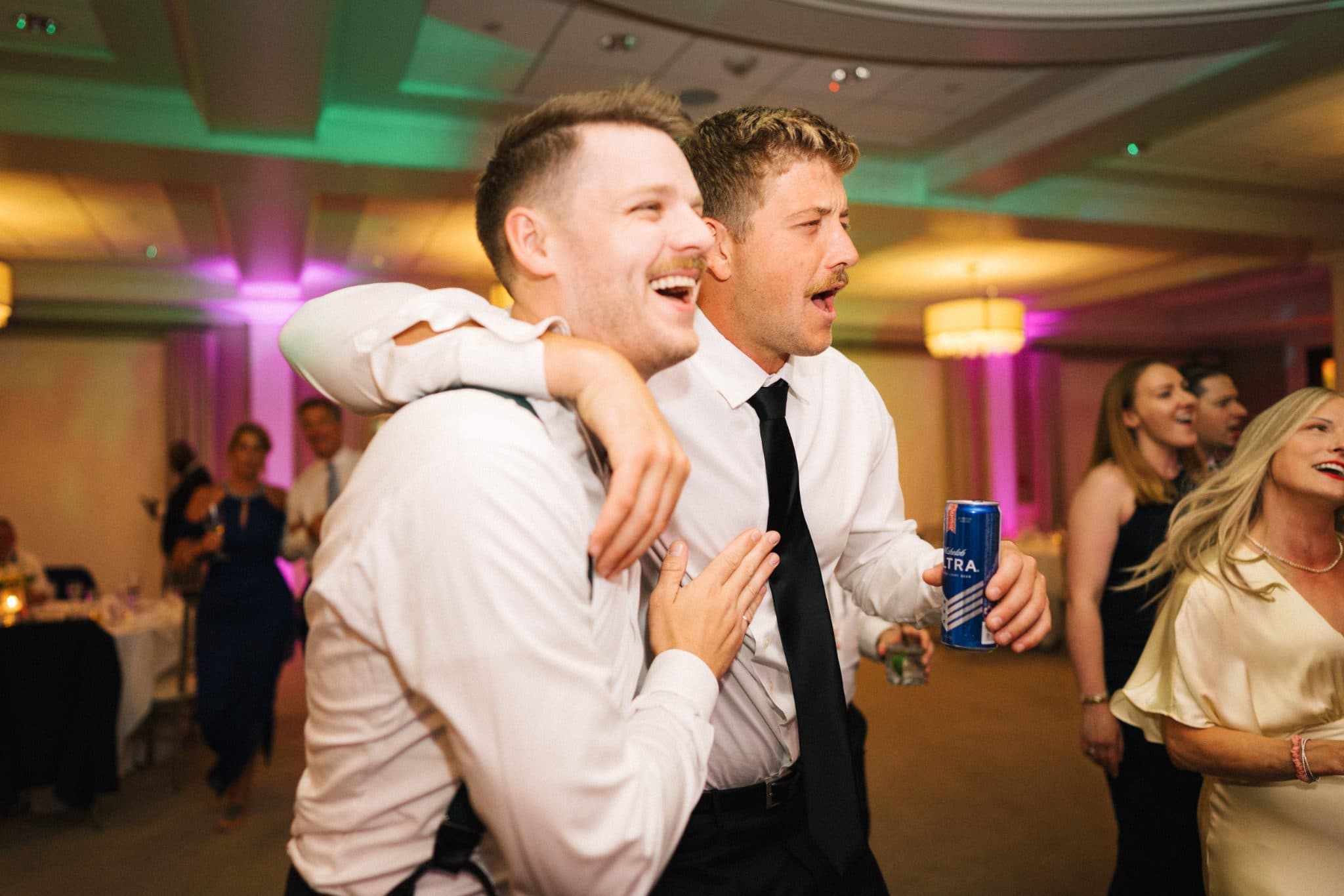 Two men in white shirts and ties are dancing together at a lively indoor event with colorful lighting.