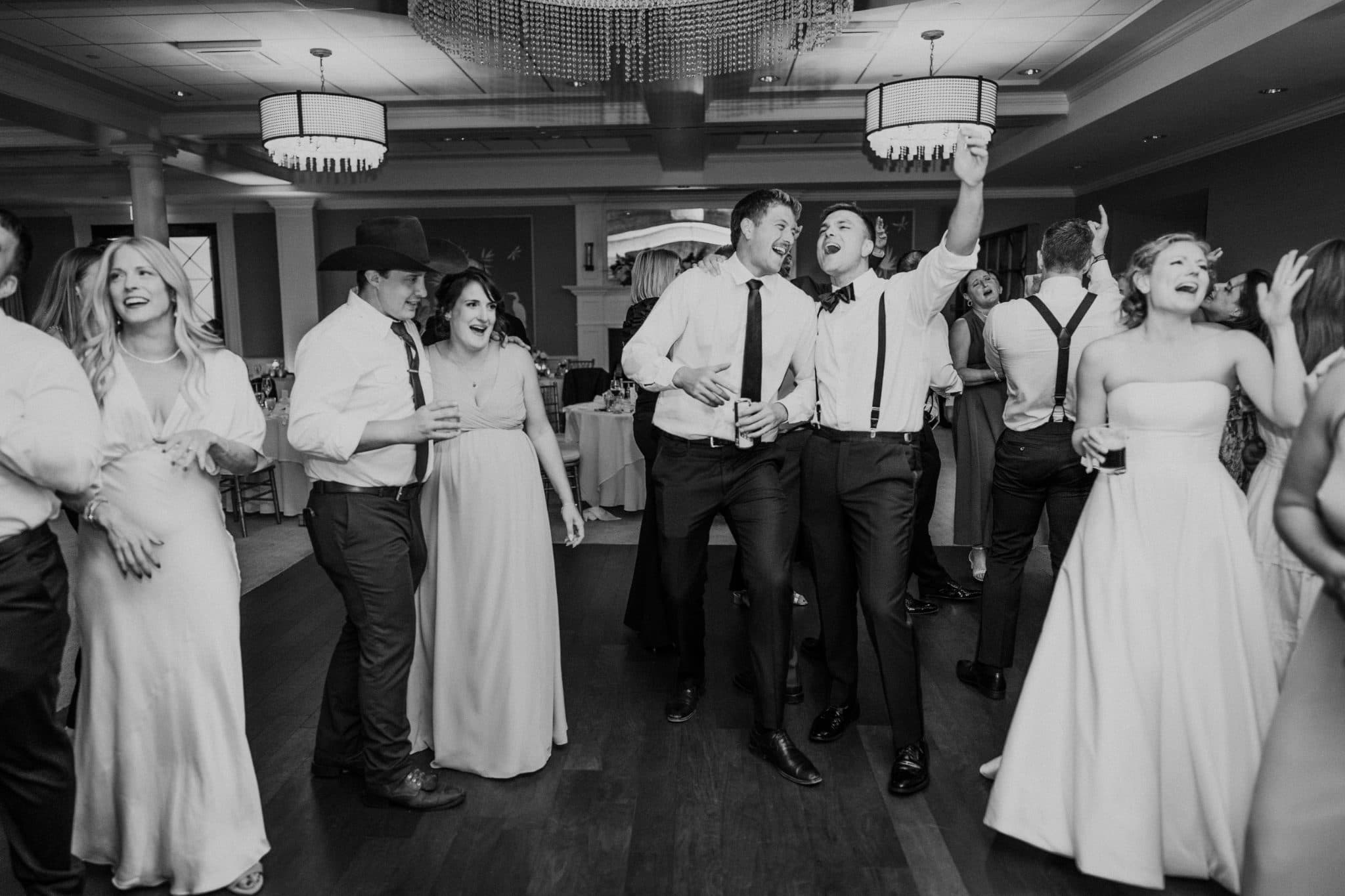 A group of people in formal attire dances and celebrates on a dance floor in a well-lit room.
