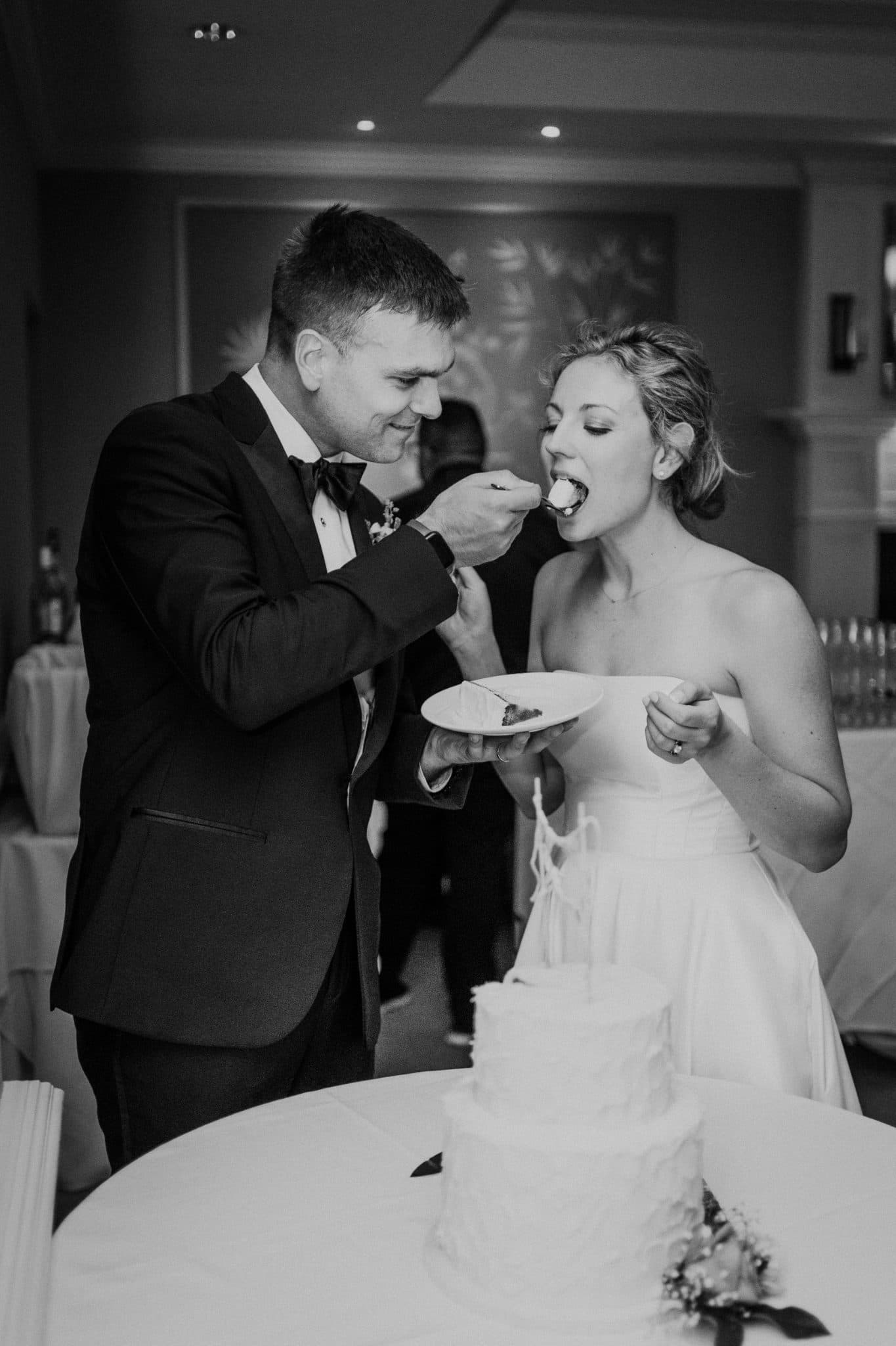A man in a suit feeds a woman in a wedding dress a piece of cake at a table.