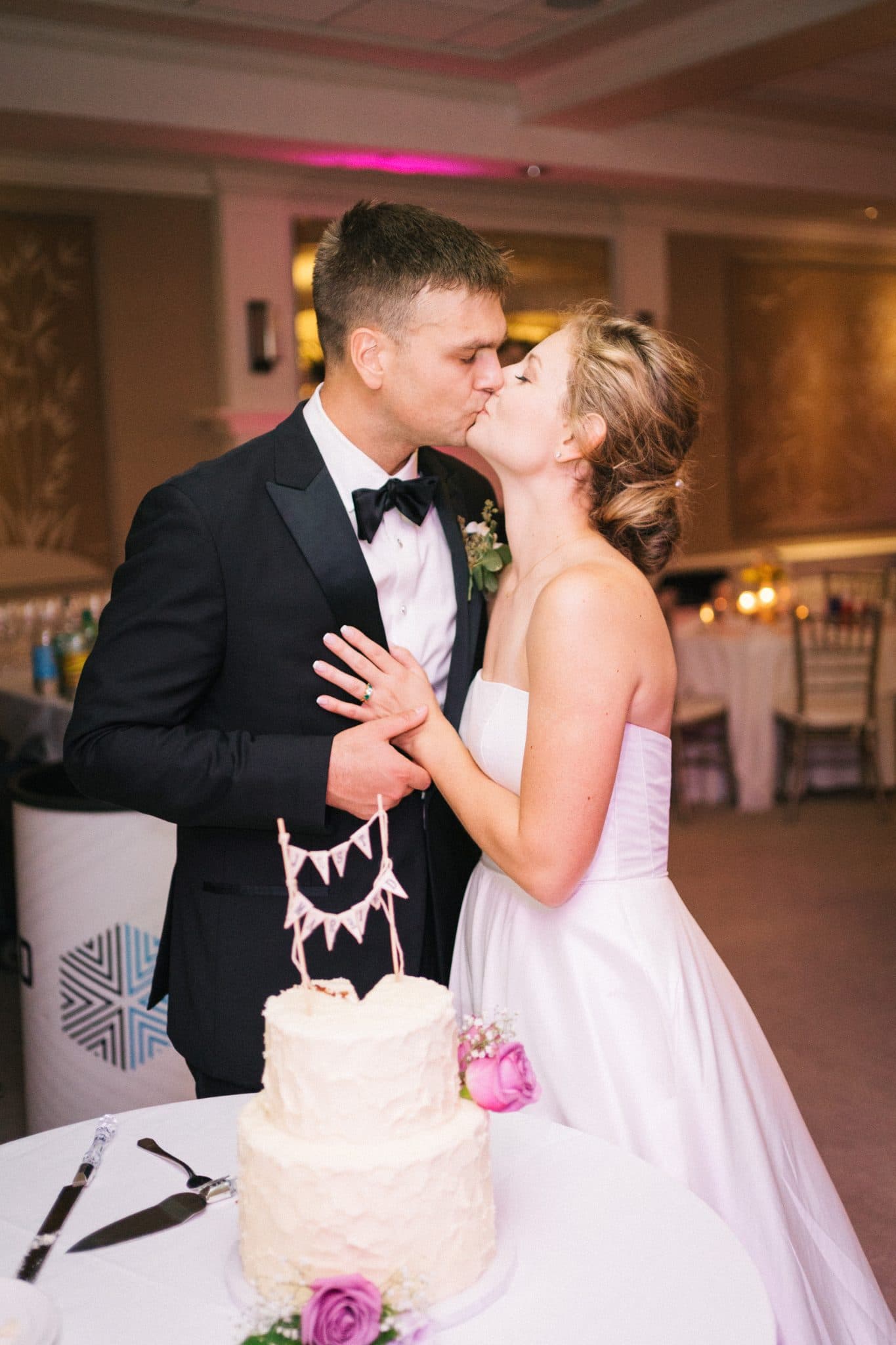 A bride and groom share a kiss beside a tiered wedding cake adorned with pink flowers.