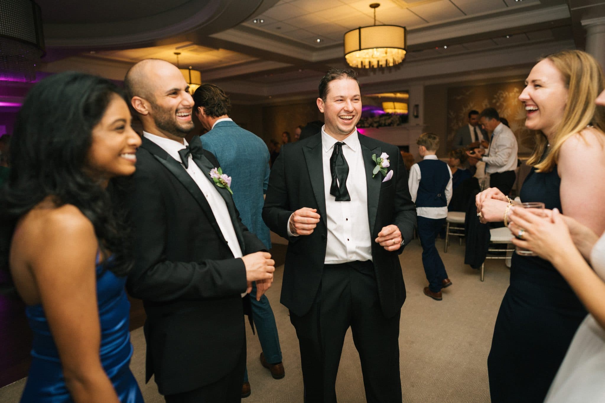 A group of people in formal attire smiles and dances in a warmly lit room.