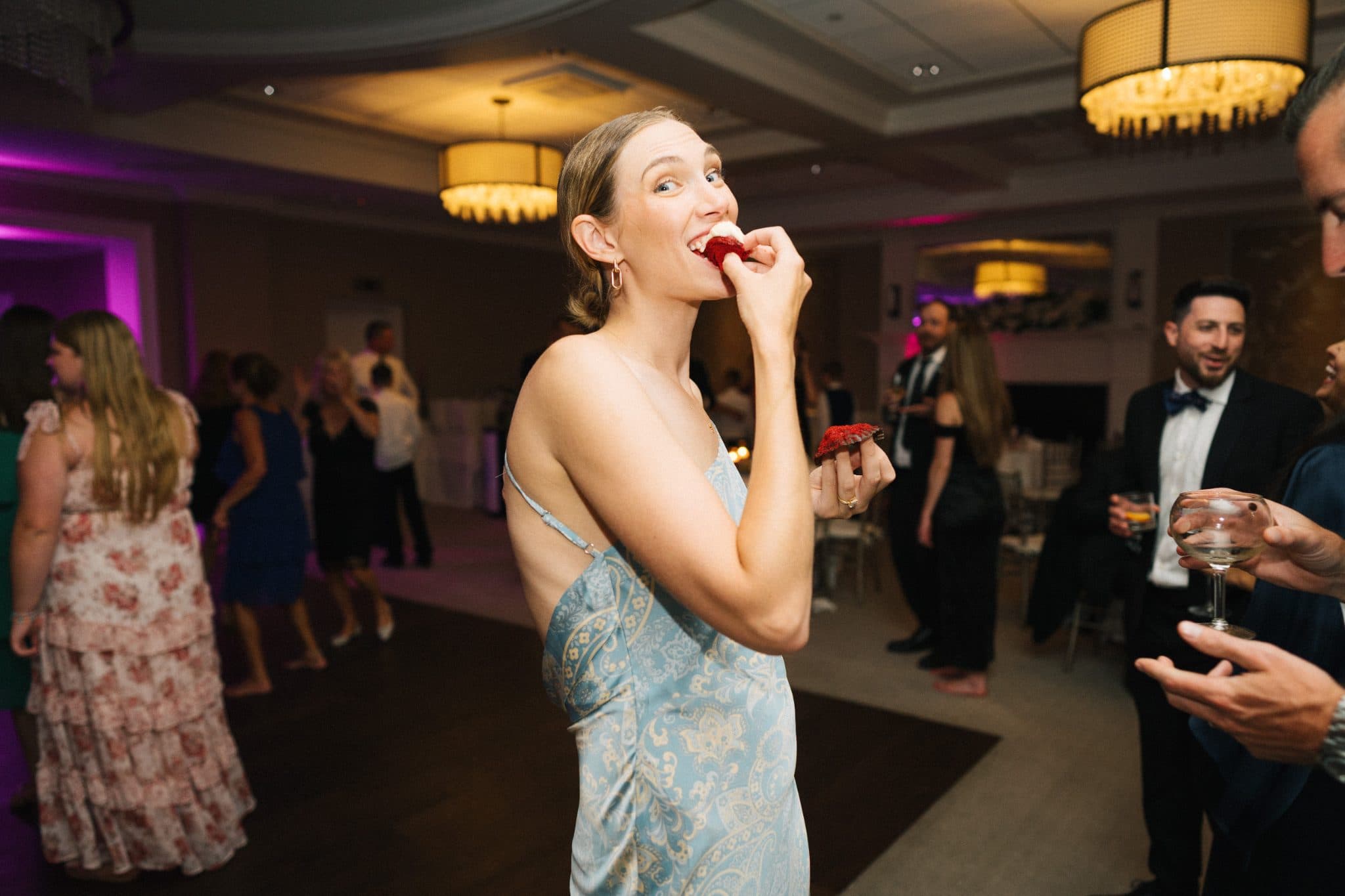 A woman in a blue dress eats a strawberry at a lively wedding reception.