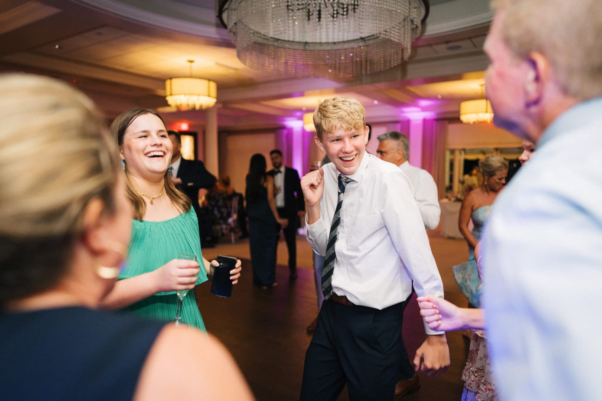 A lively wedding reception scene with guests dancing and socializing under chandeliers.