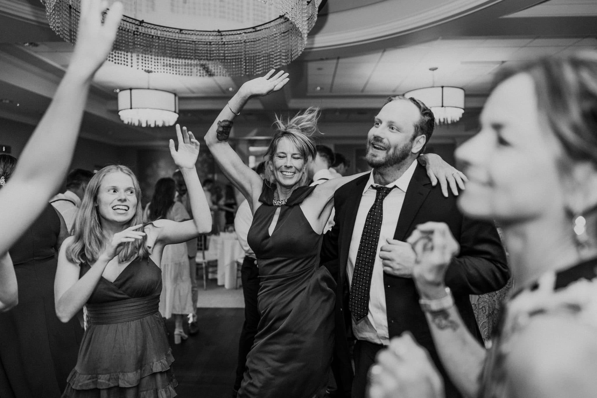 A group of people dances joyfully in a room with chandeliers, wearing formal attire.