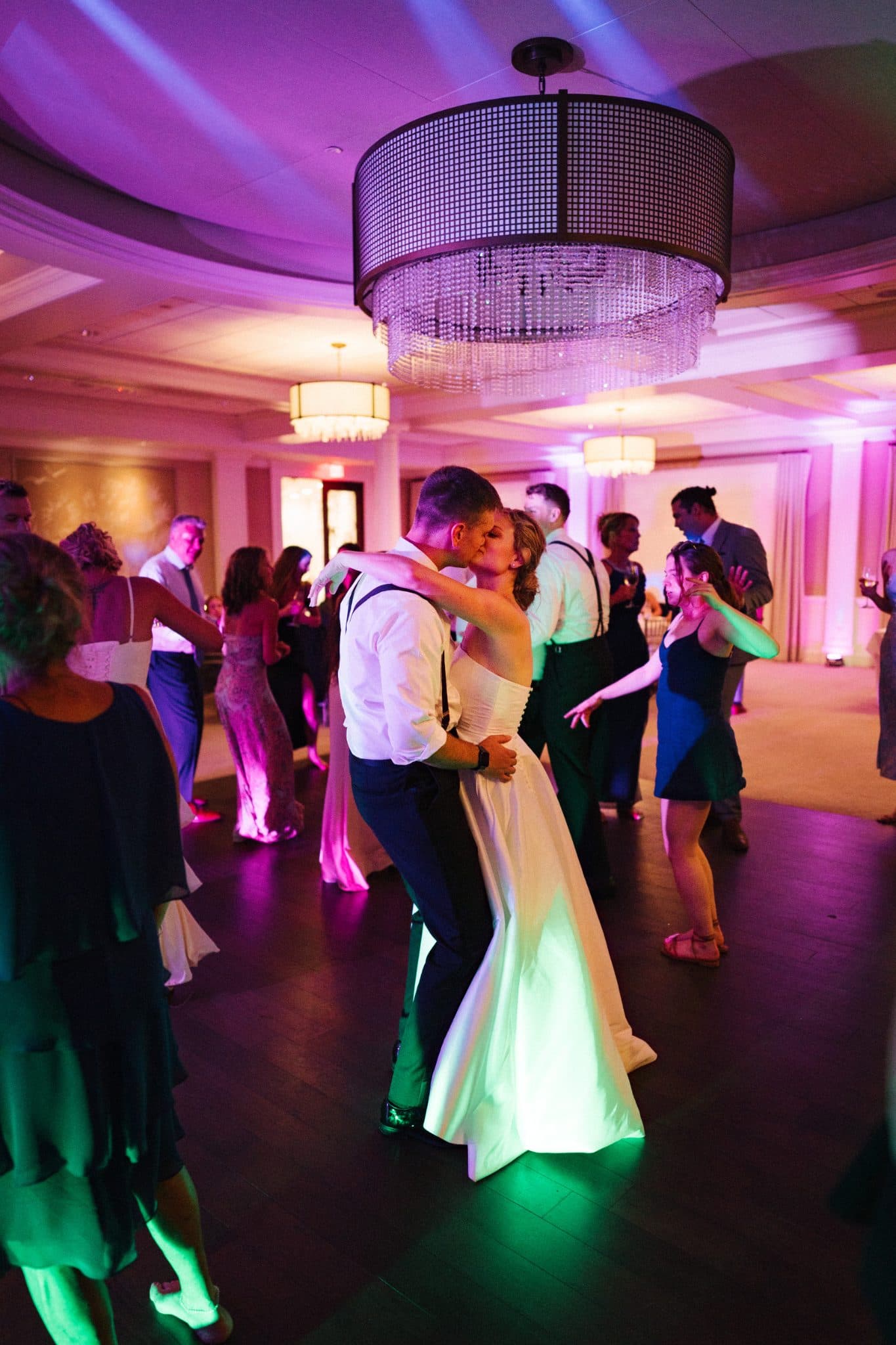 A bride and groom share a dance under a chandelier, surrounded by guests in a warmly lit room.