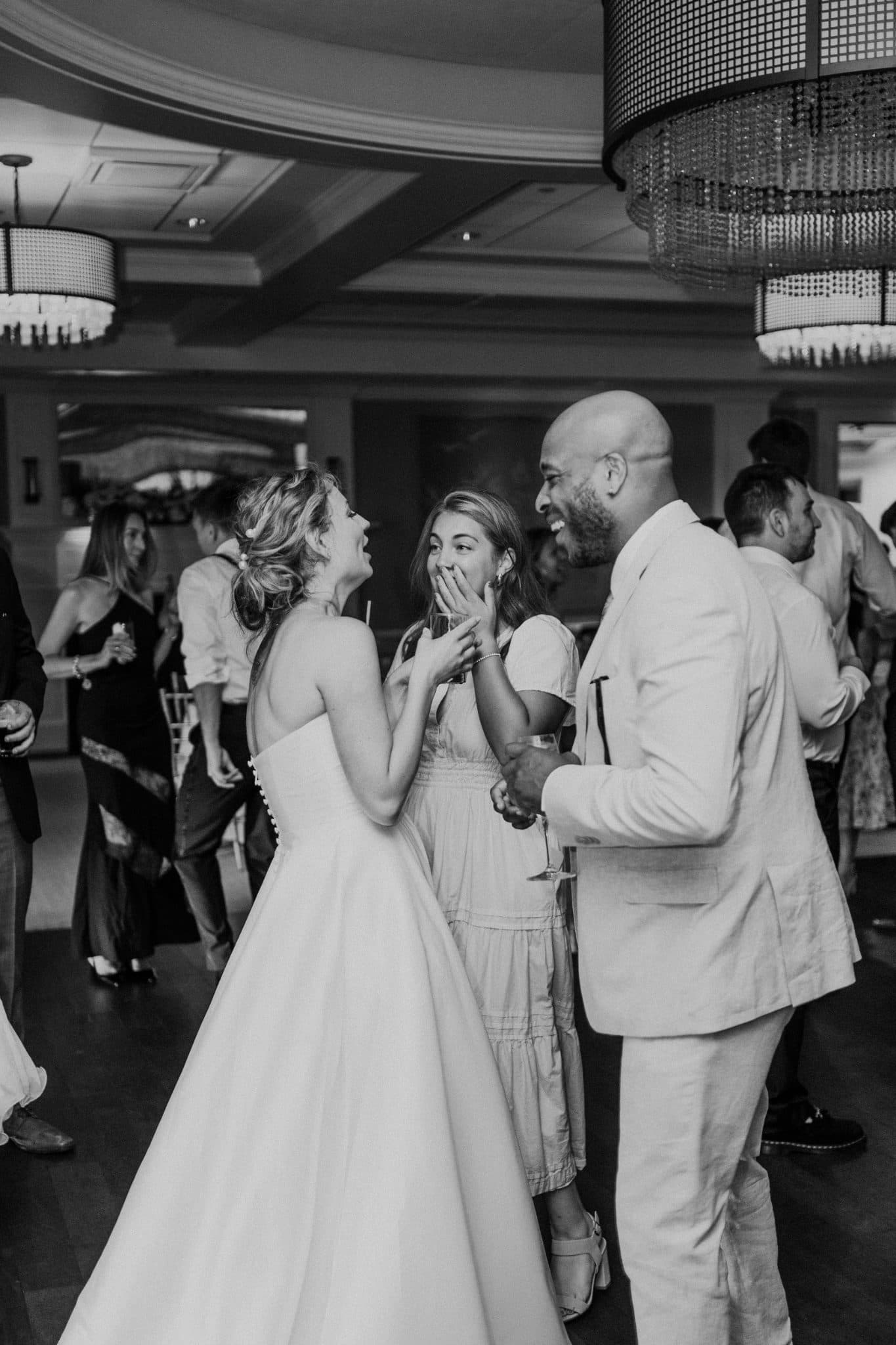 A bride in a white gown dances with two guests in a well-lit indoor venue.