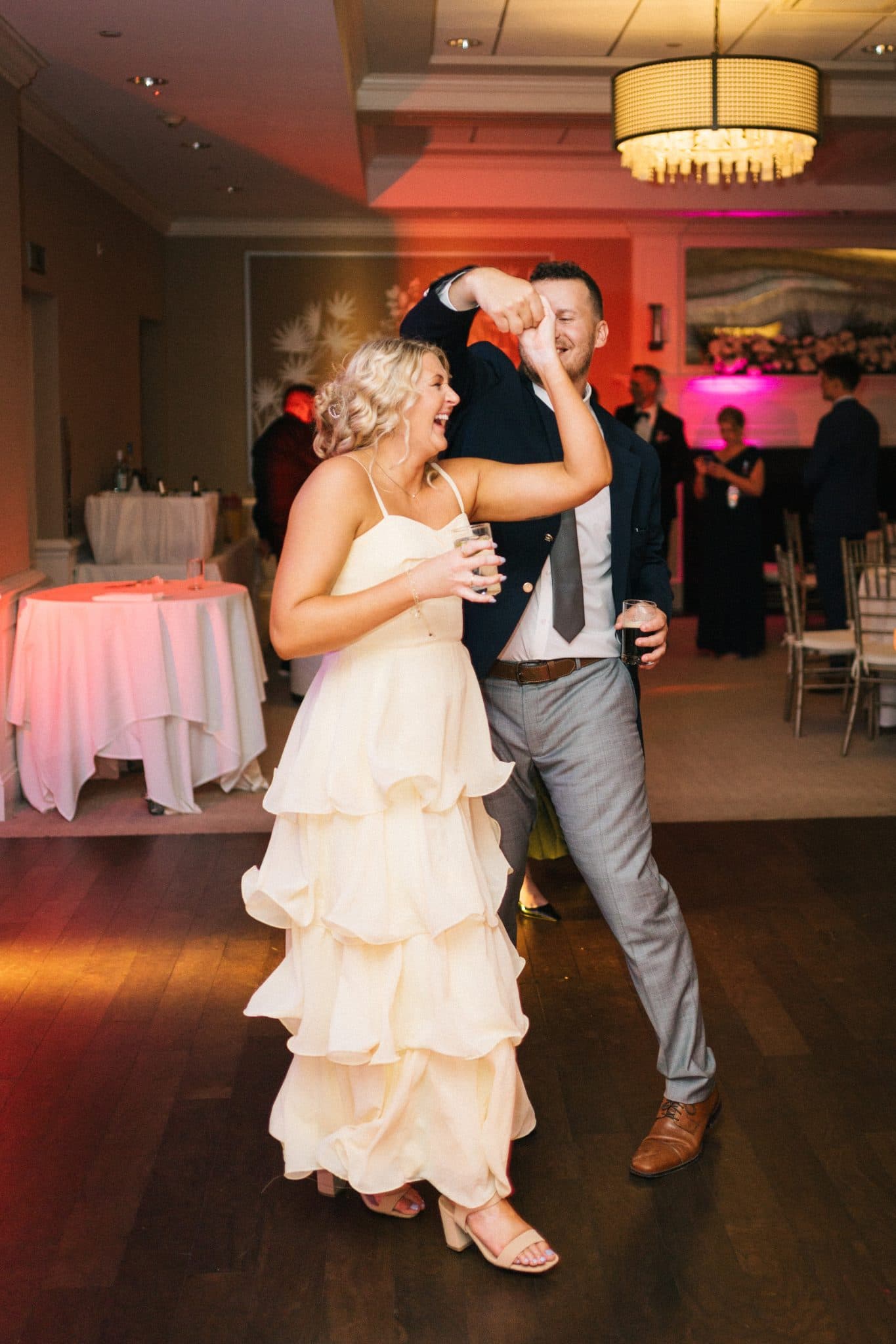 A couple dances joyfully on a wooden floor, holding drinks, with a warmly lit room and tables in the background.