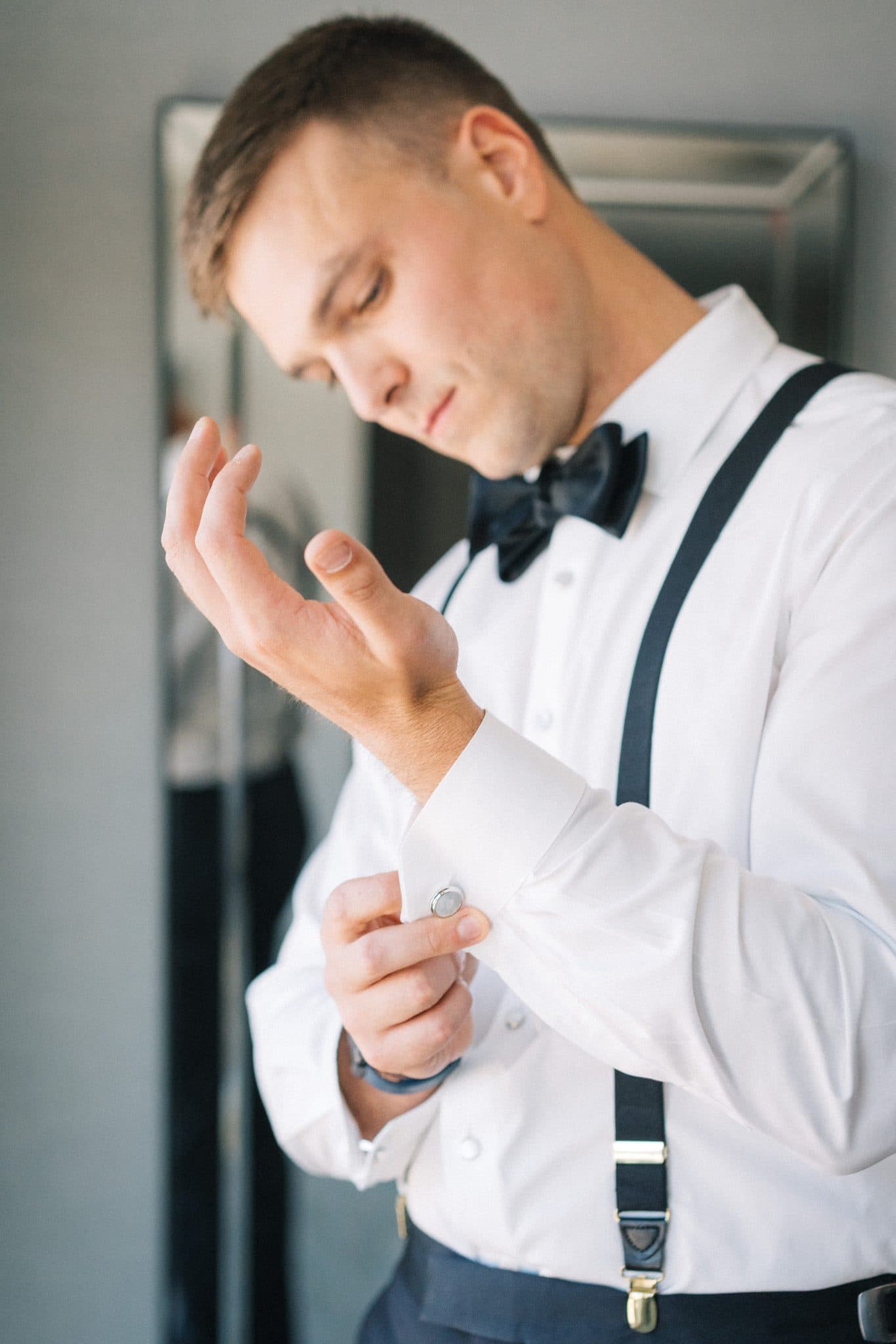 A man adjusts his white shirt cuff while wearing a black bow tie and suspenders.