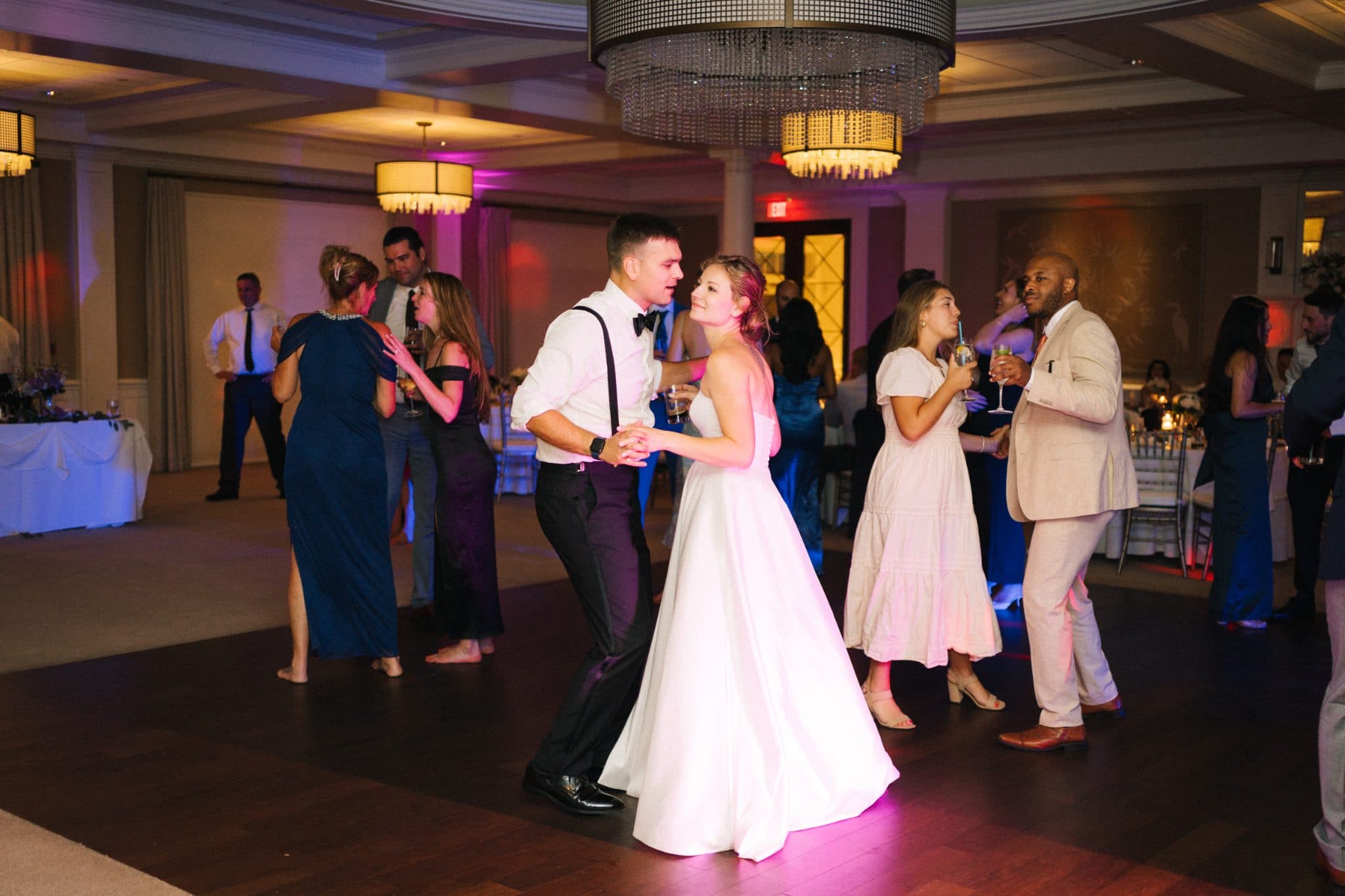A bride in a white dress dances with a man in a white suit at a wedding reception with guests in formal attire.