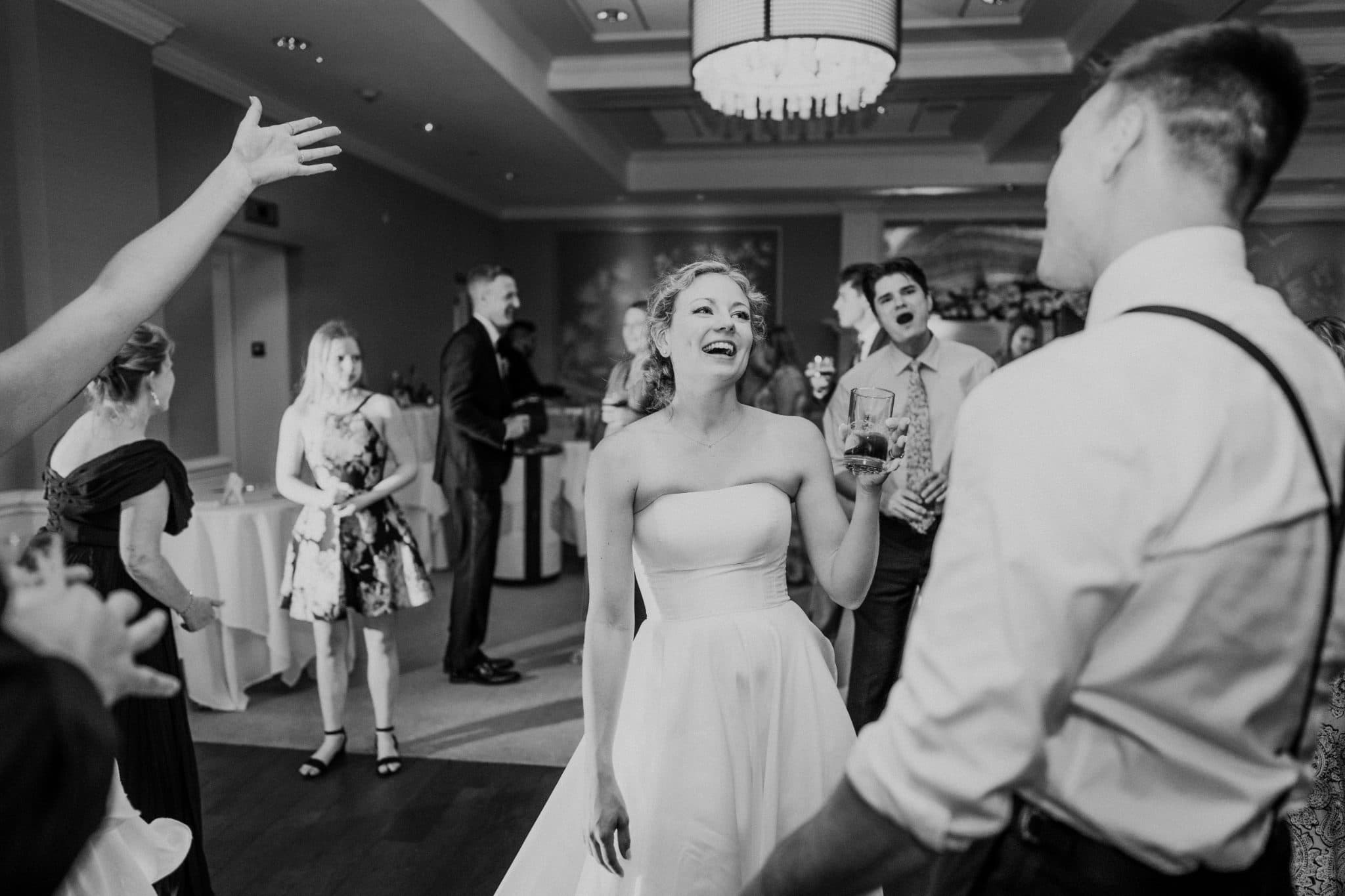 A bride in a strapless dress dances joyfully at a wedding reception, surrounded by guests in formal attire.