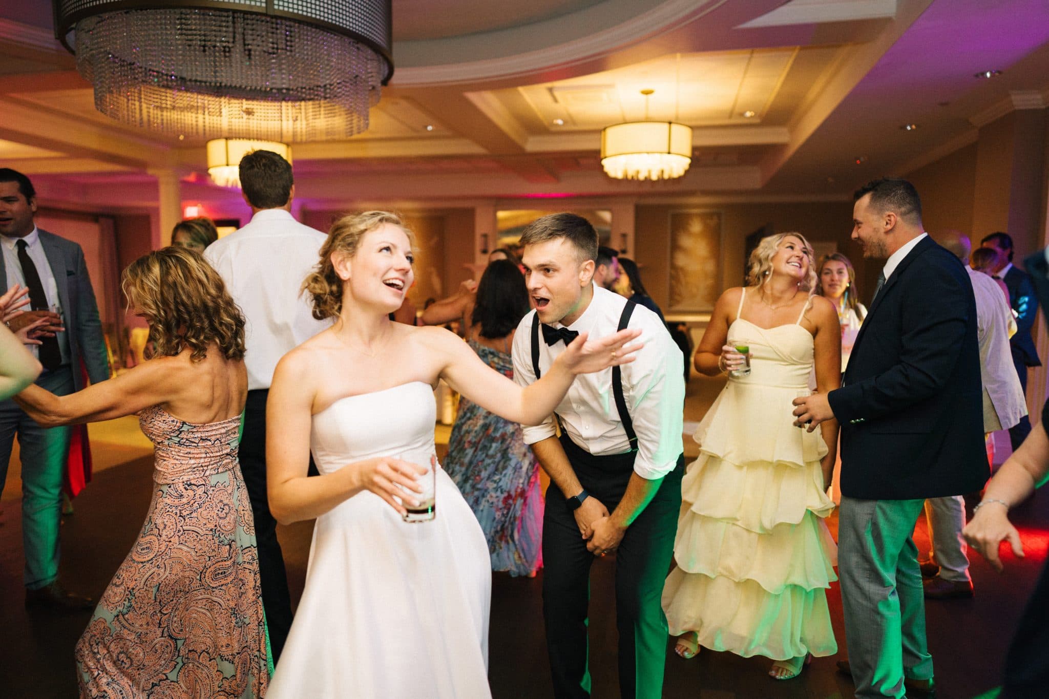 A bride in a white dress dances with guests in a lively, well-lit room.