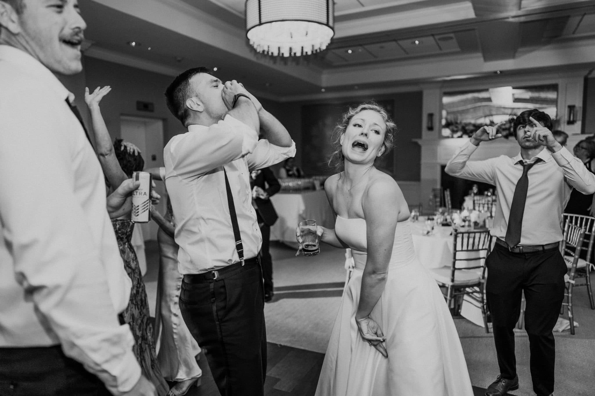 A bride and groom dance with guests at a wedding reception, with one guest holding a drink and another making a playful gesture.