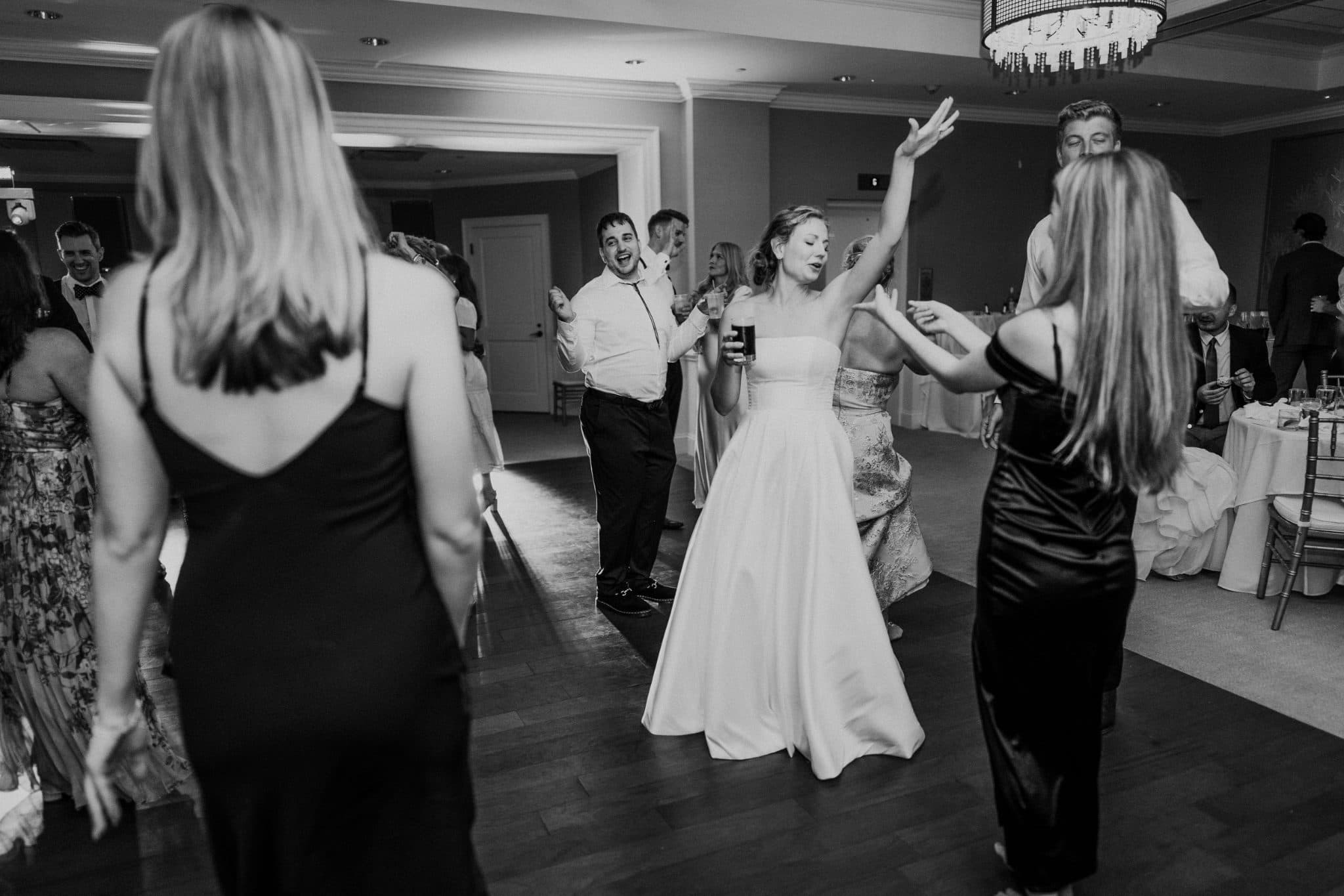A bride in a white dress dances joyfully on a wooden floor surrounded by guests in a warmly lit room.