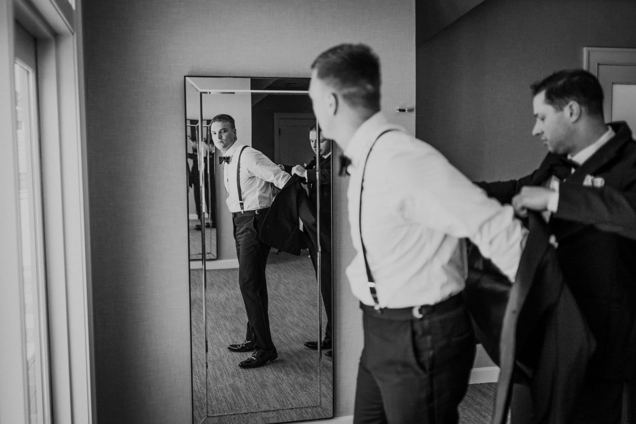 A man adjusts his bow tie in front of a mirror while another person assists.