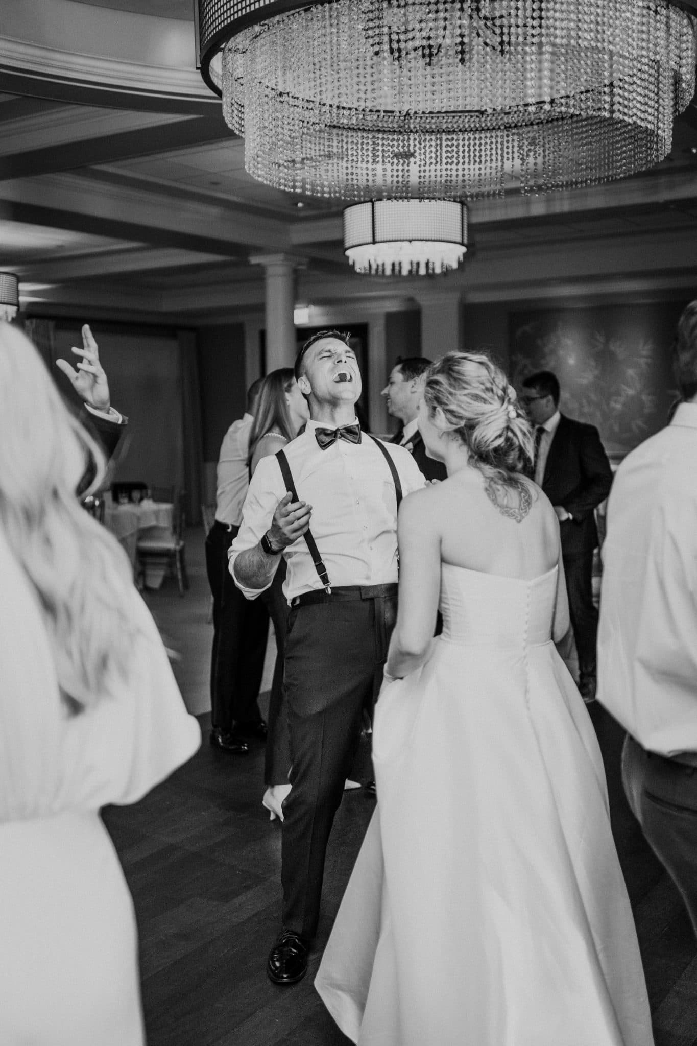A bride in a white dress dances with a man in a bow tie and suspenders under a large chandelier in a room with other guests.
