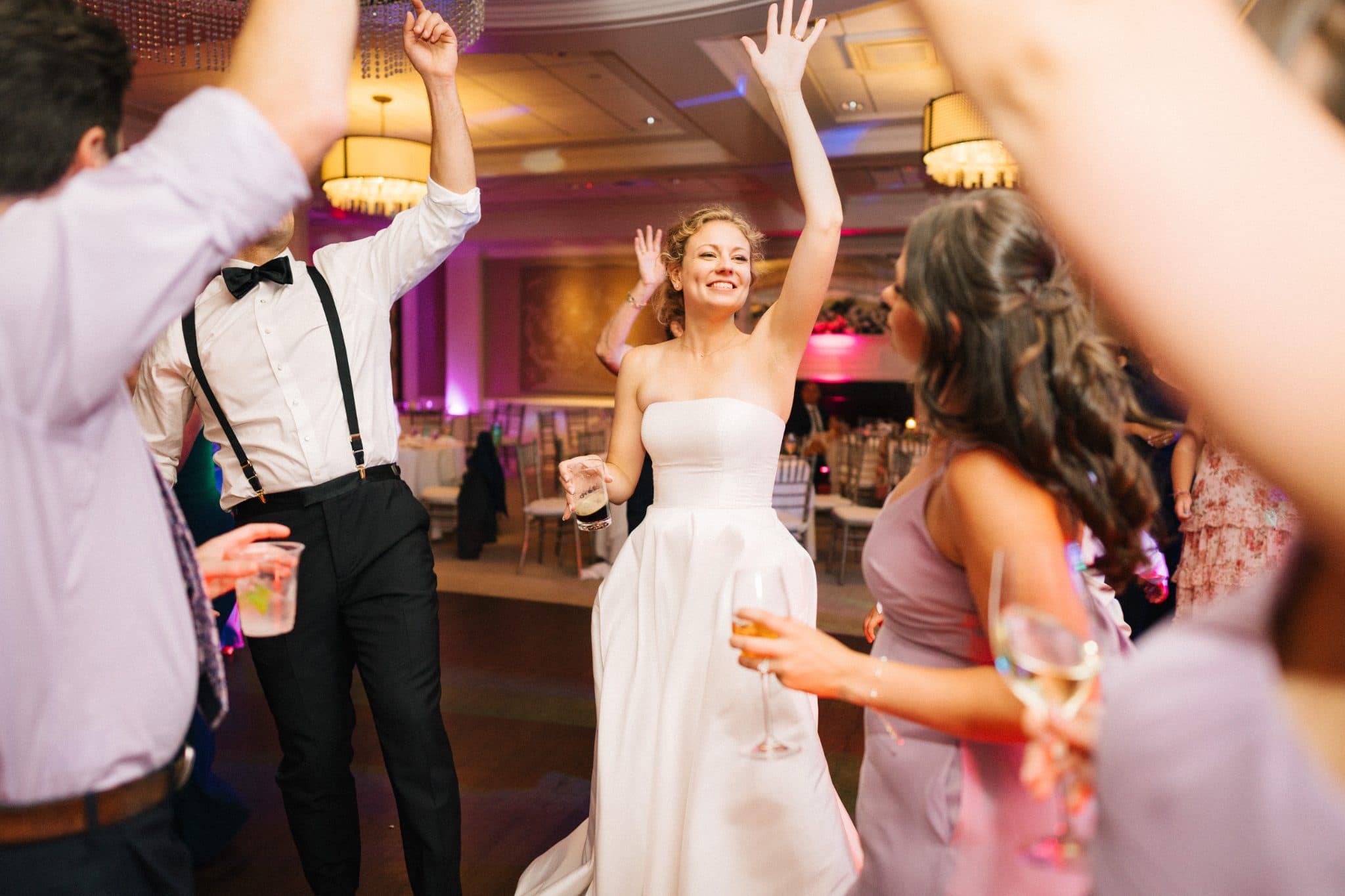 A bride in a white dress dances joyfully with guests in a brightly lit room.