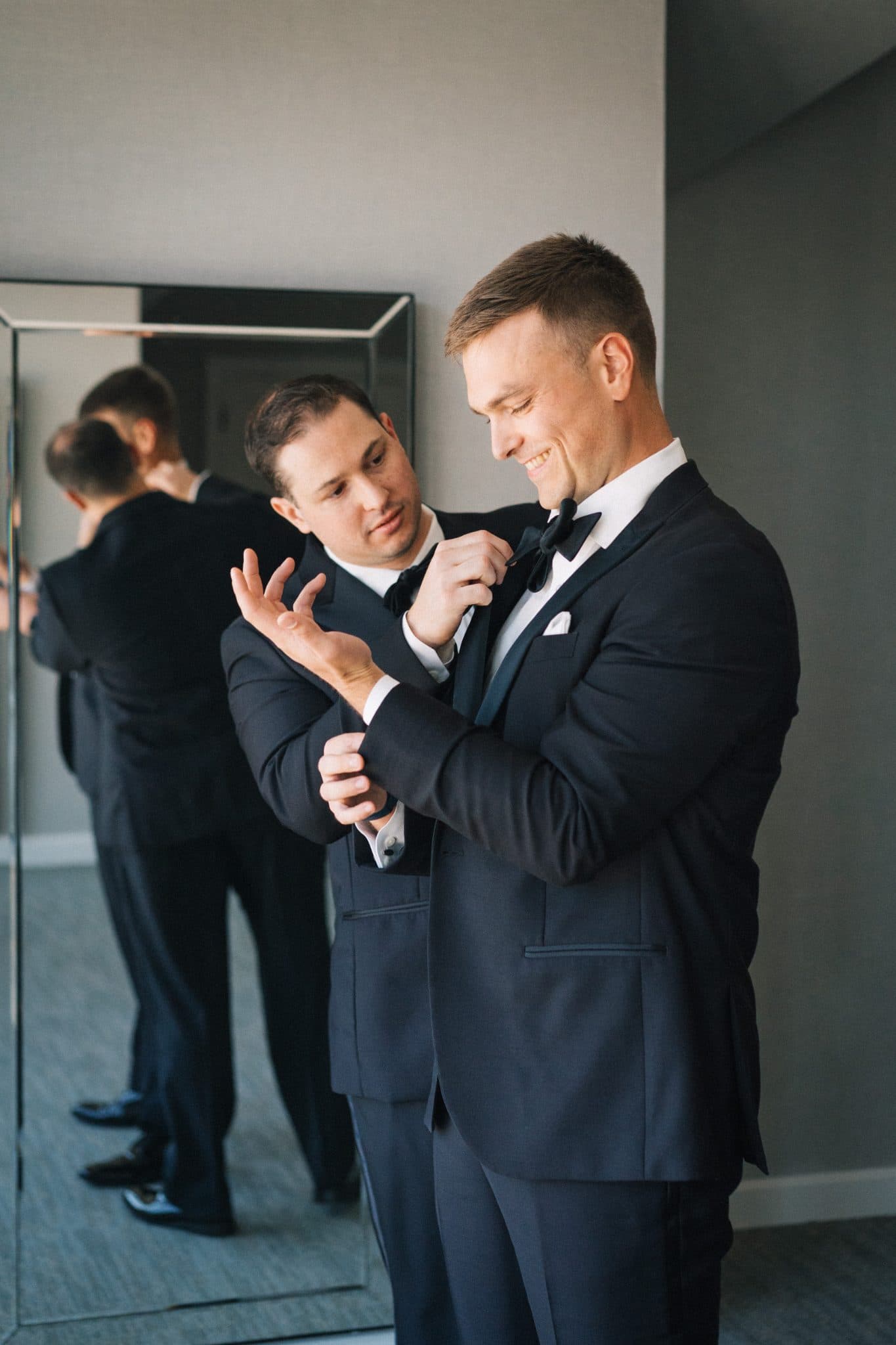 A man in a suit adjusts his tie while another man assists, both reflected in a mirror.