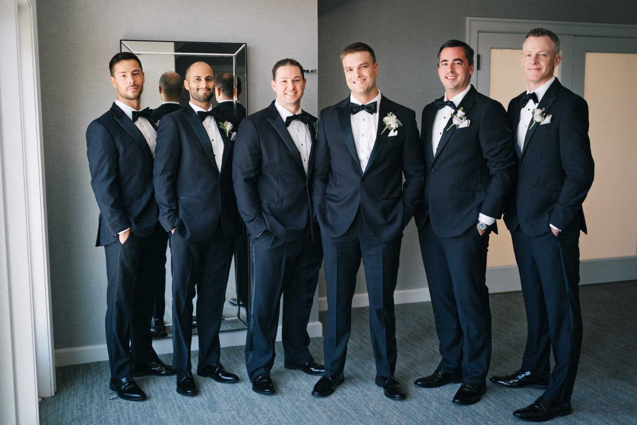 Six men in formal black tuxedos stand together indoors, smiling and posing for a photograph.