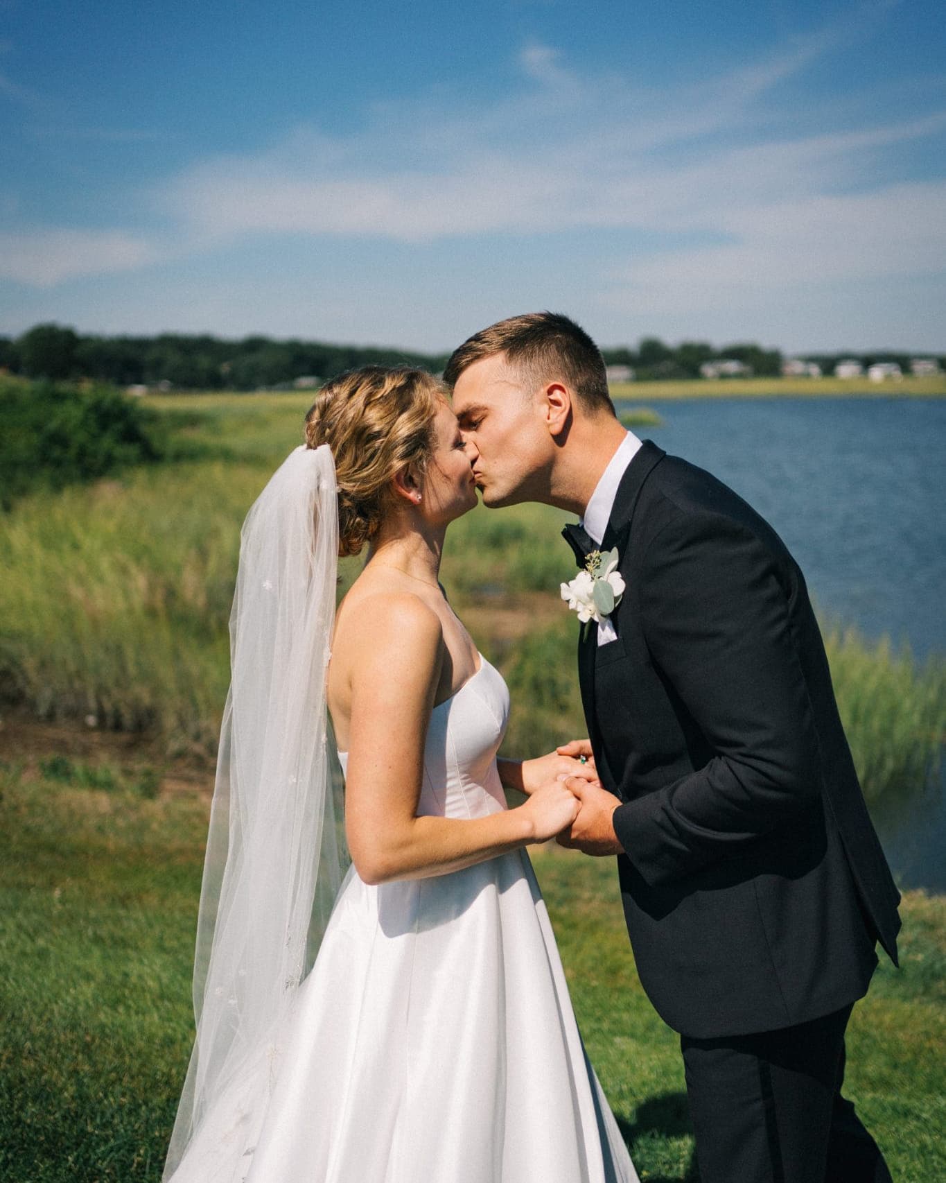 A bride in a white dress and veil kisses a groom in a dark suit by a body of water under a blue sky.