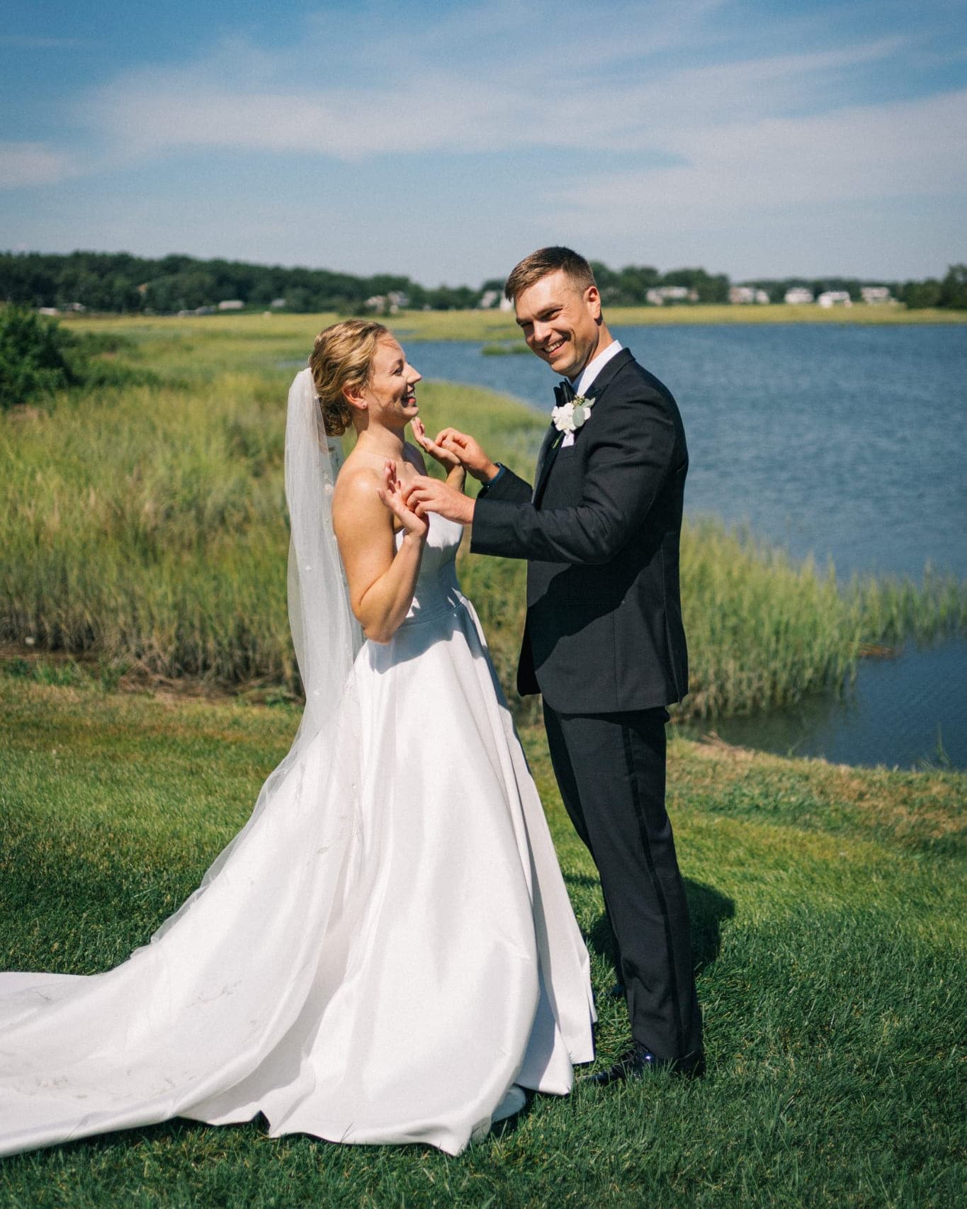 A bride in a white wedding dress and a groom in a dark suit stand on grass near a body of water under a blue sky.