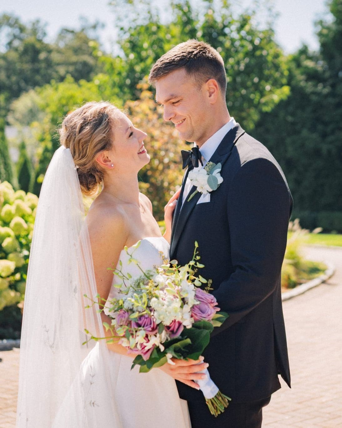 A bride in a white dress and veil holds a bouquet of flowers, standing next to a man in a dark suit with a bow tie, both smiling and looking at each…