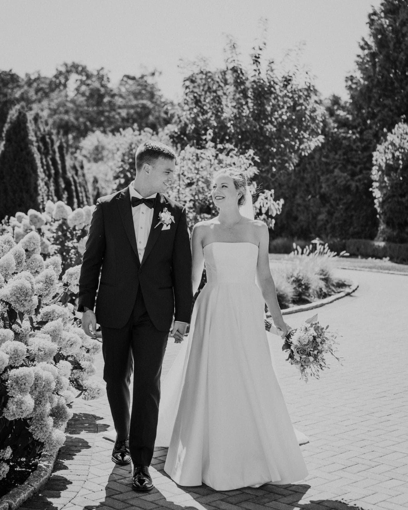 A bride in a strapless gown and a groom in a suit walk hand in hand on a brick path surrounded by lush greenery.