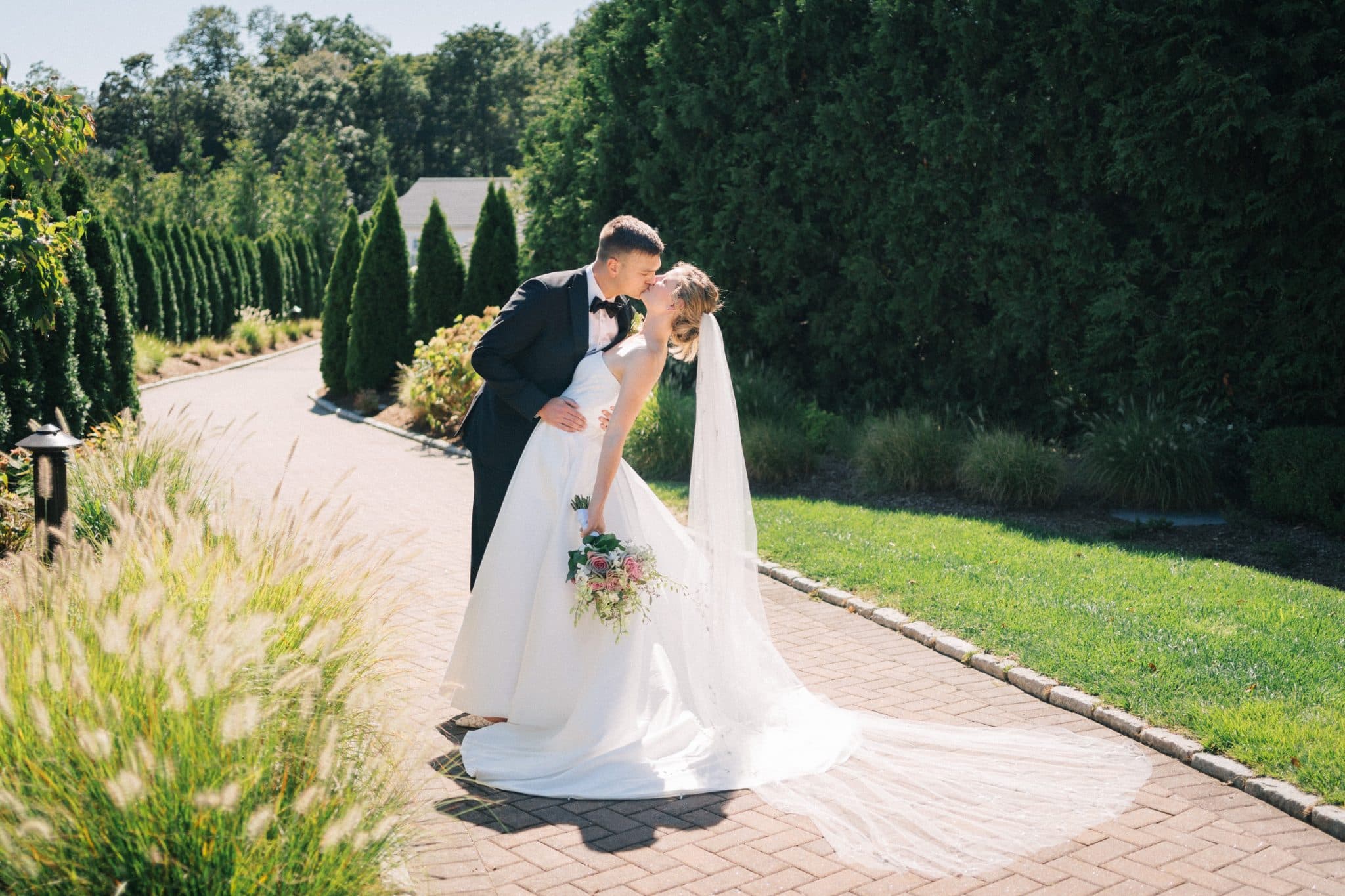 A bride in a white gown and veil kisses a groom in a black suit on a brick pathway surrounded by greenery.