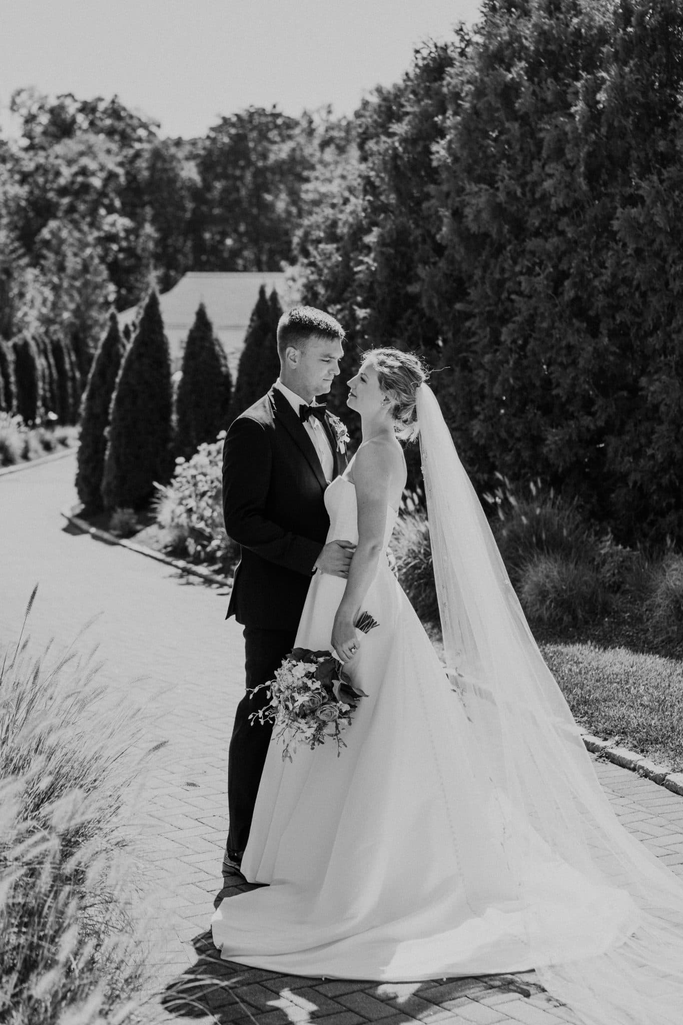 A bride in a white gown and veil stands beside a groom in a black suit, holding hands, with a backdrop of trees and a brick path.
