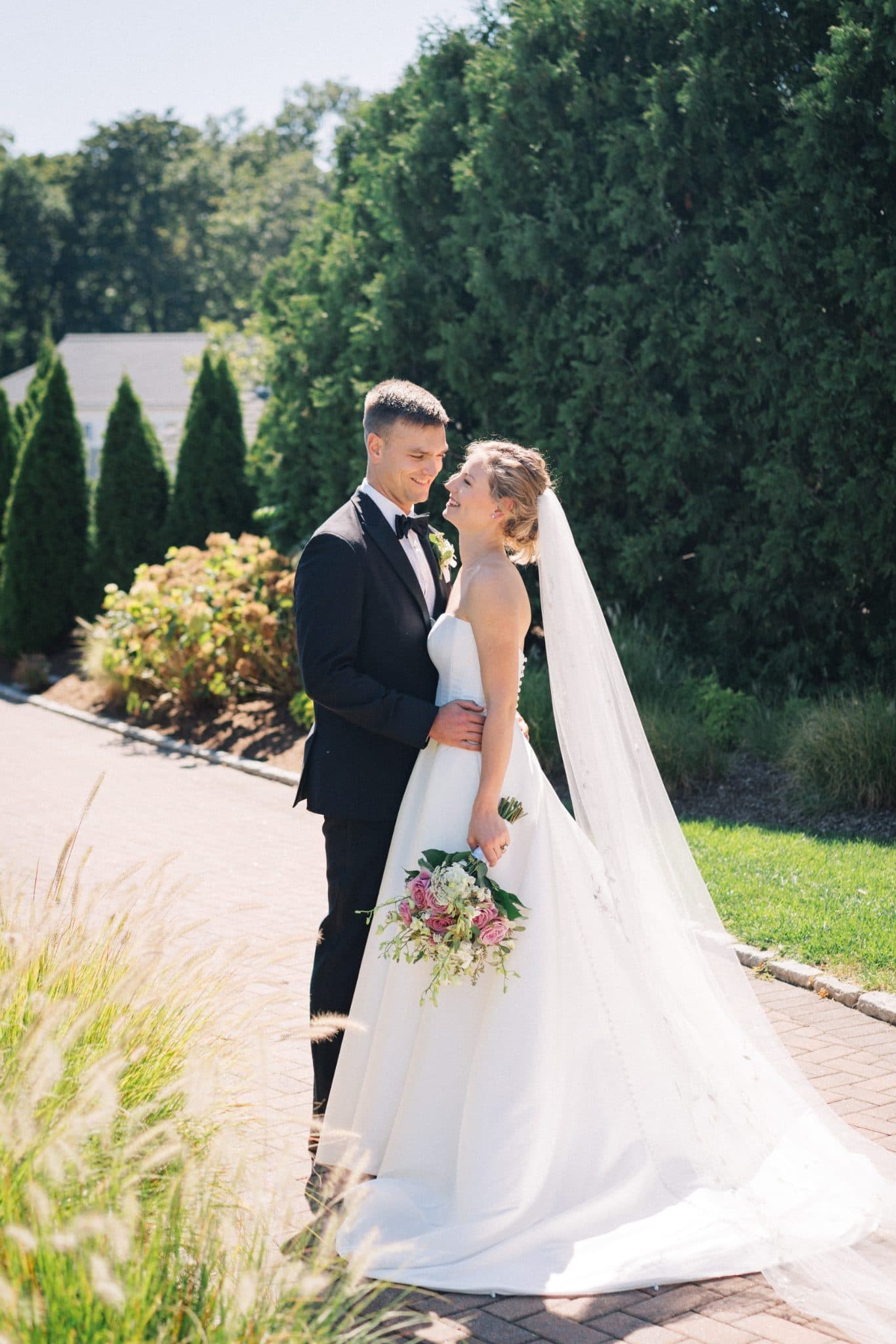 A bride in a white gown and veil holds a bouquet, embracing a groom in a black suit against a backdrop of greenery and a brick path.