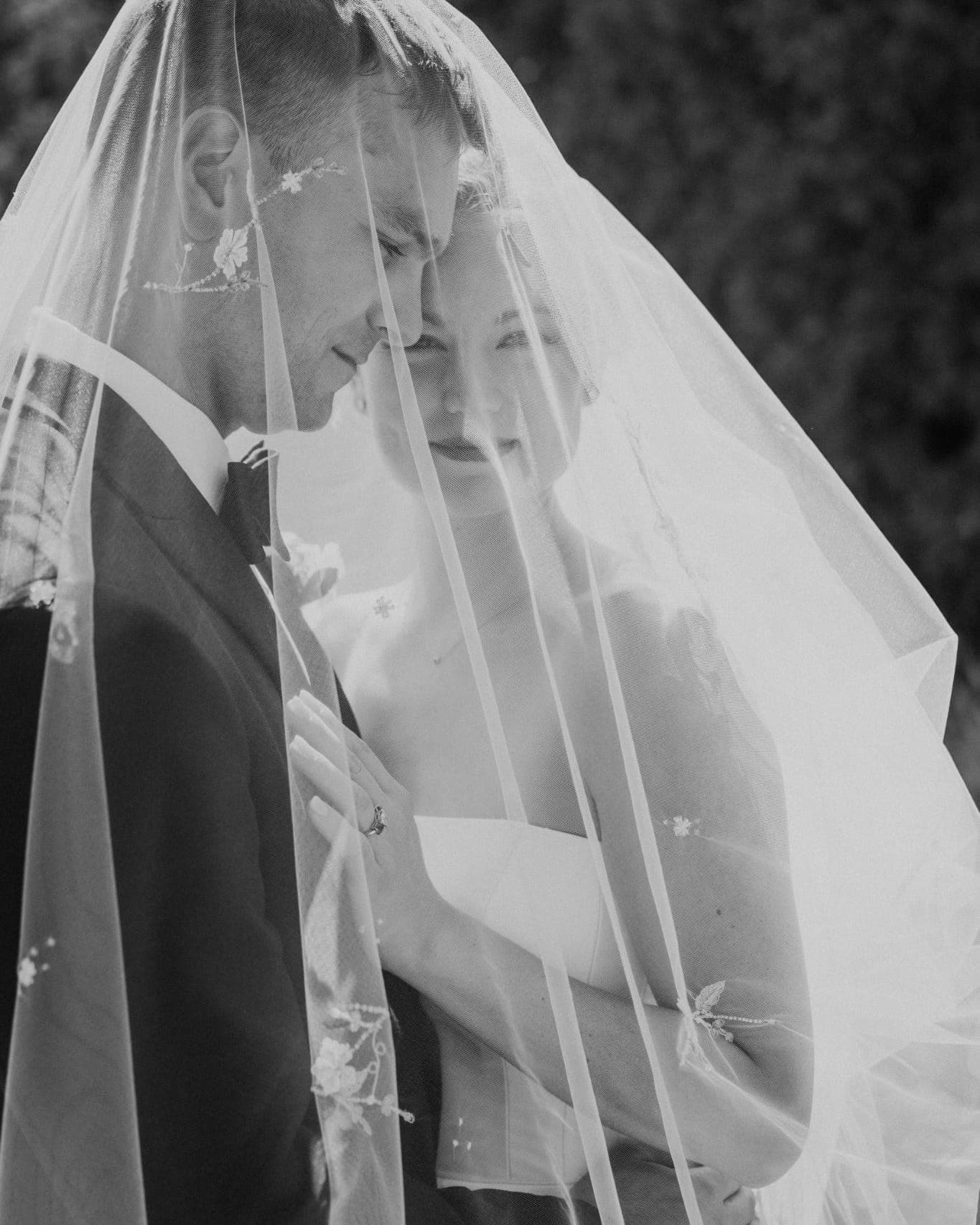 A bride and groom stand close under a veil, the bride's hand on the groom's chest.