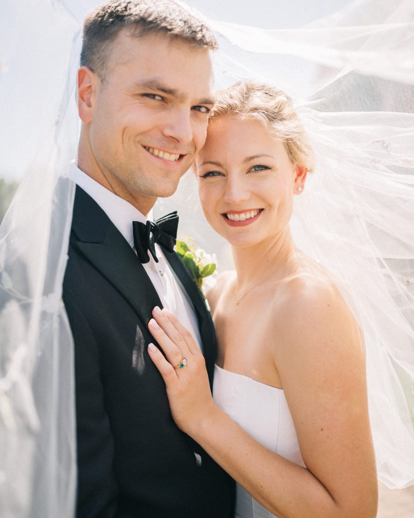 A bride in a white strapless dress and a groom in a black tuxedo pose together, with the bride's hand resting on the groom's chest.