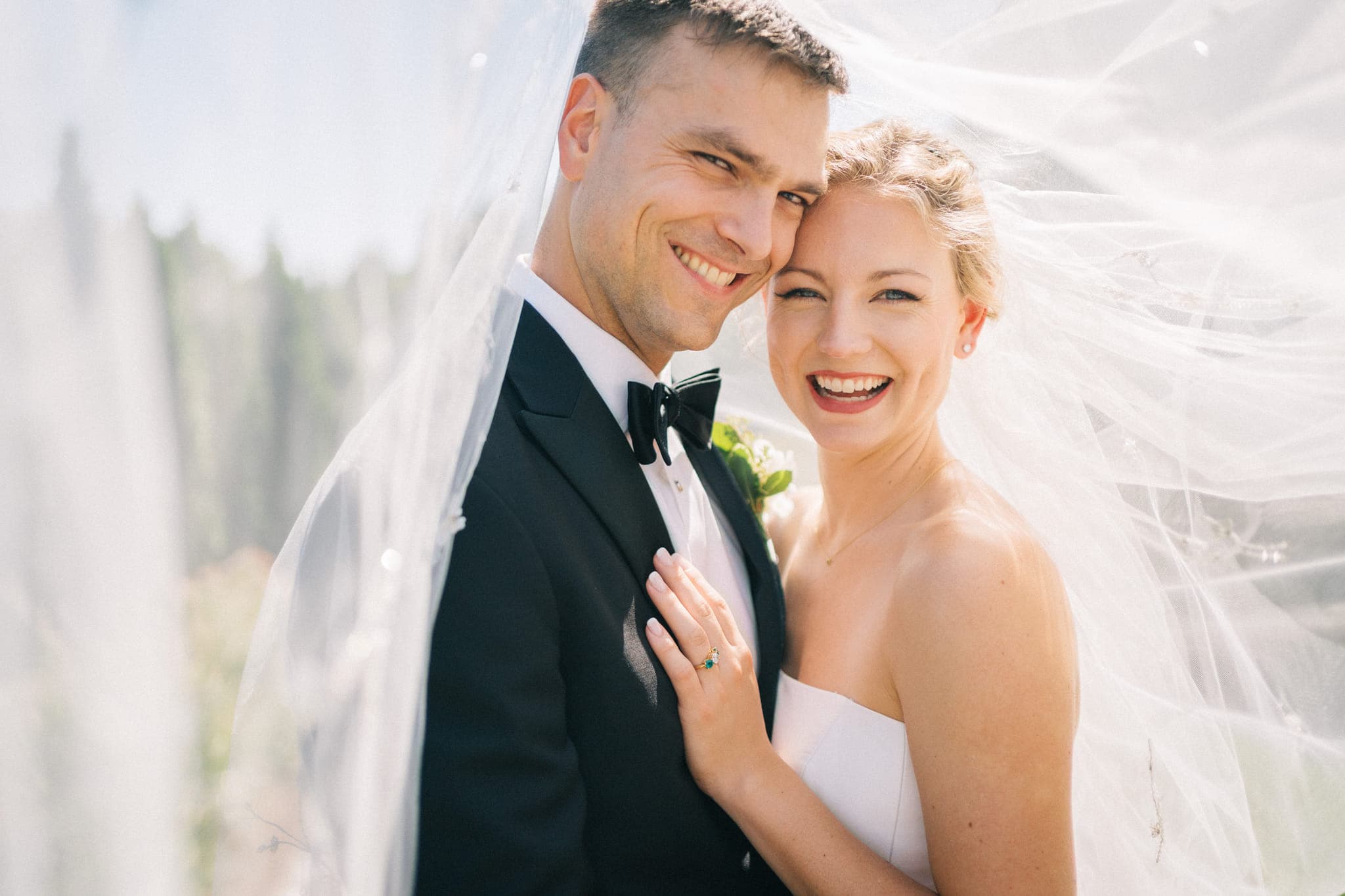A bride in a strapless white dress and a groom in a black tuxedo smile under a flowing white veil.