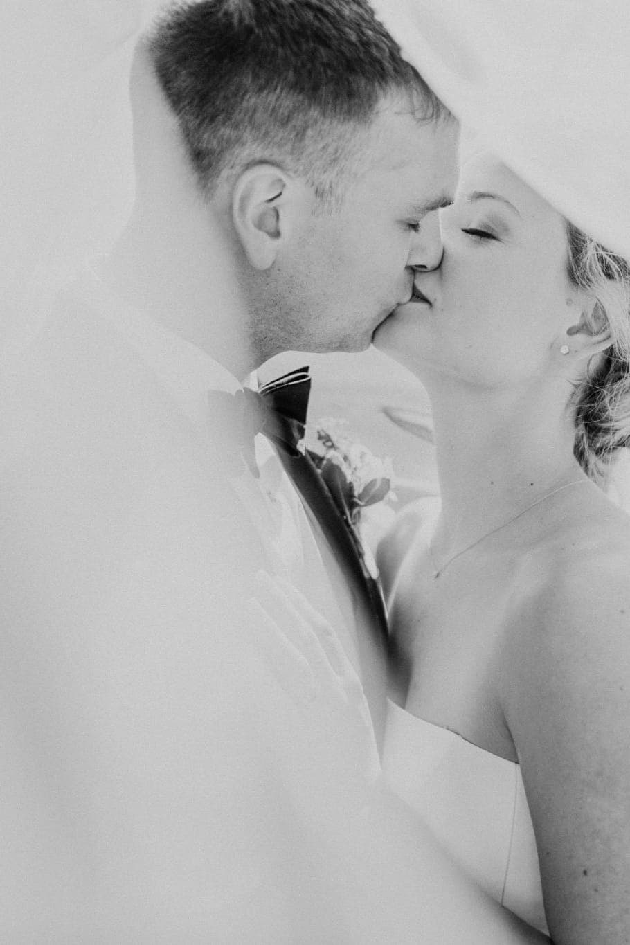 A bride and groom share a close kiss under a veil, both dressed formally.
