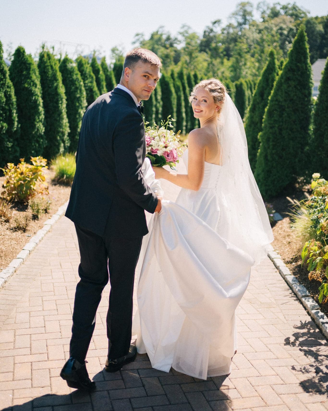 A bride in a white gown and veil holds a bouquet, while a groom in a dark suit supports her dress on a brick path surrounded by greenery.