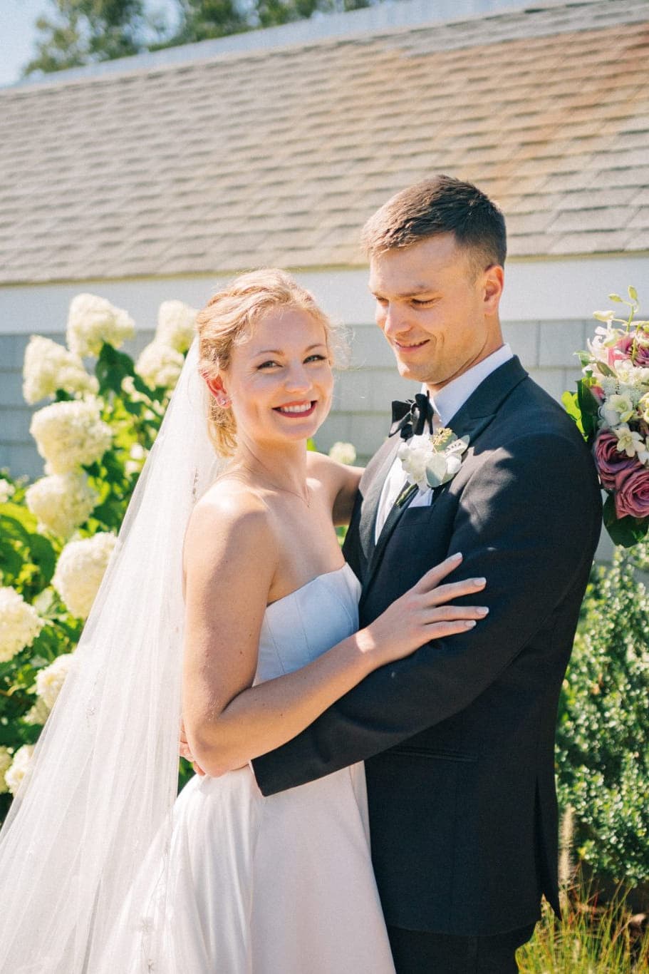 A bride in a white dress and veil hugs a groom in a dark suit, standing in front of a house with greenery and flowers.