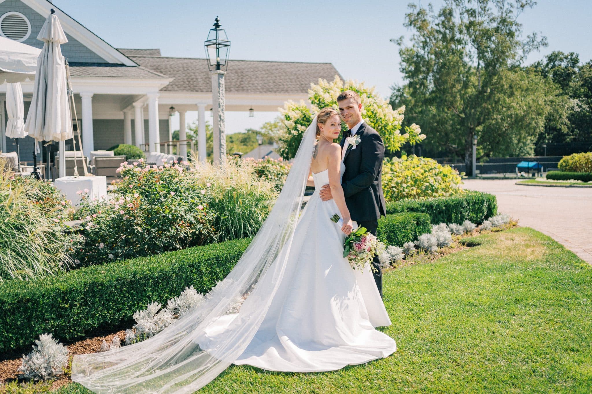 A bride in a white wedding dress with a long veil and a groom in a black suit stand on a grassy lawn with a garden and a building in the background.