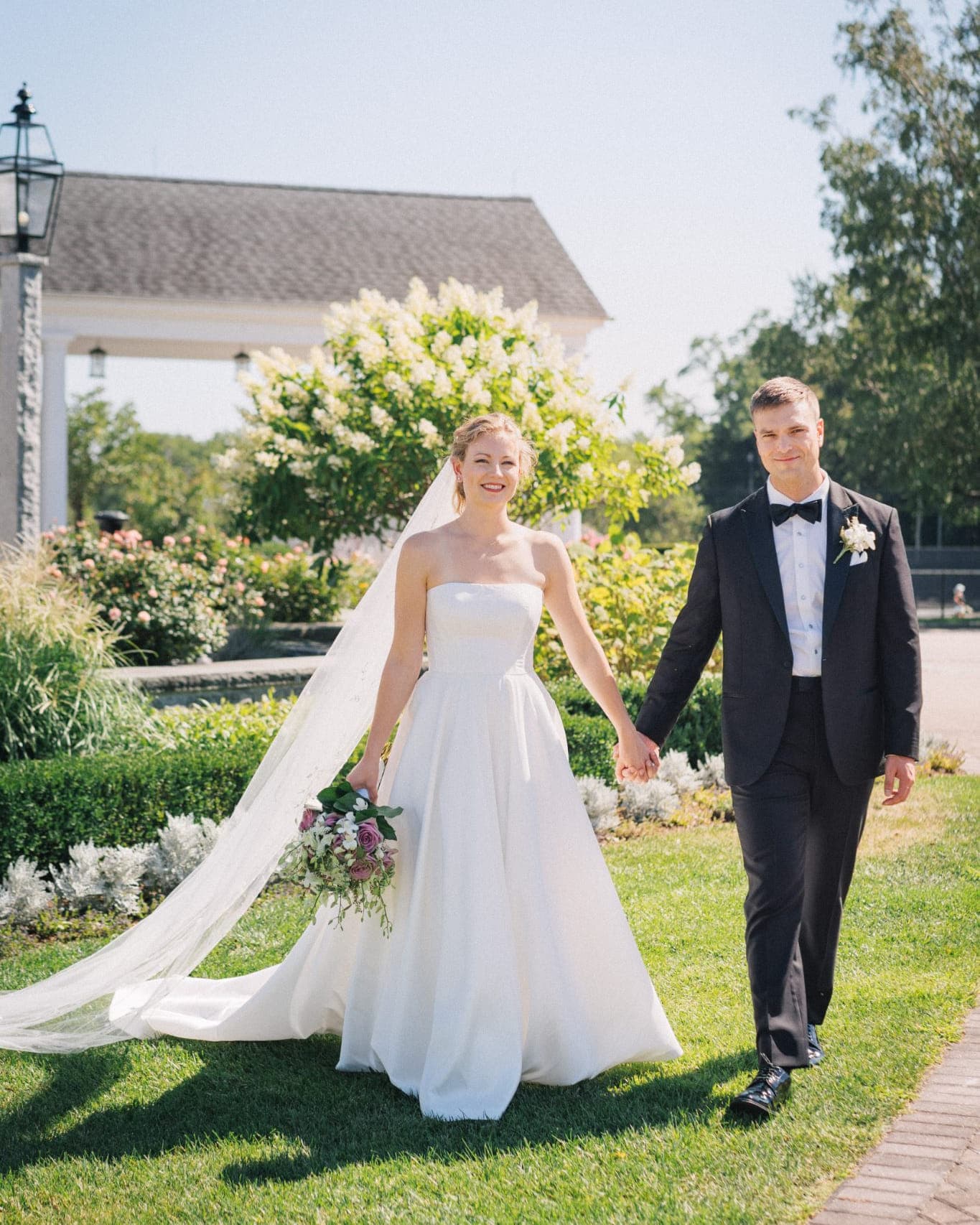 A bride in a strapless wedding gown and veil holds hands with a groom in a black suit, walking on a grassy path with a garden and building in the background.