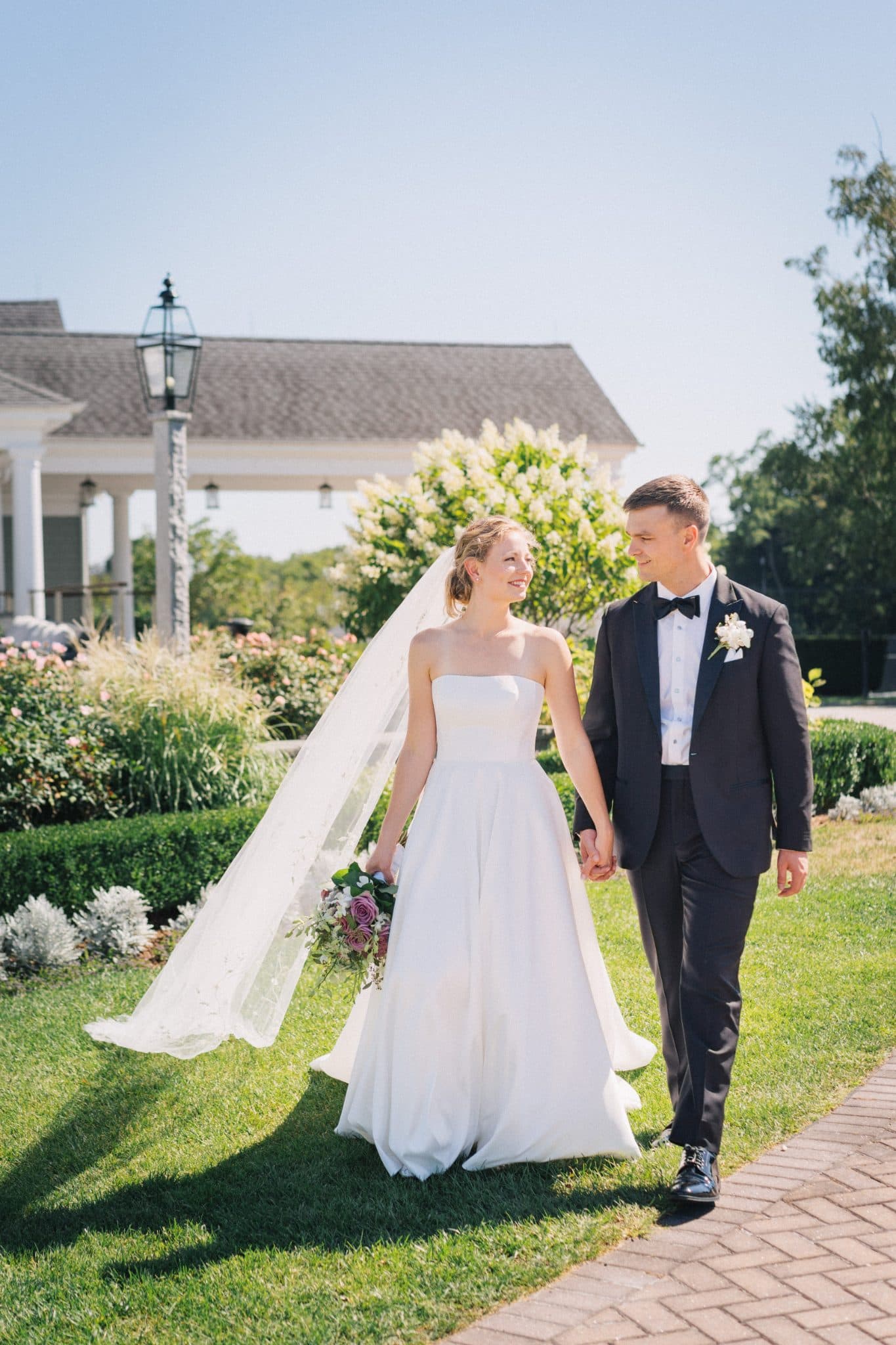 A bride in a strapless white gown and veil holds hands with a groom in a black tuxedo, walking on a grassy path near a building with a lamp post.