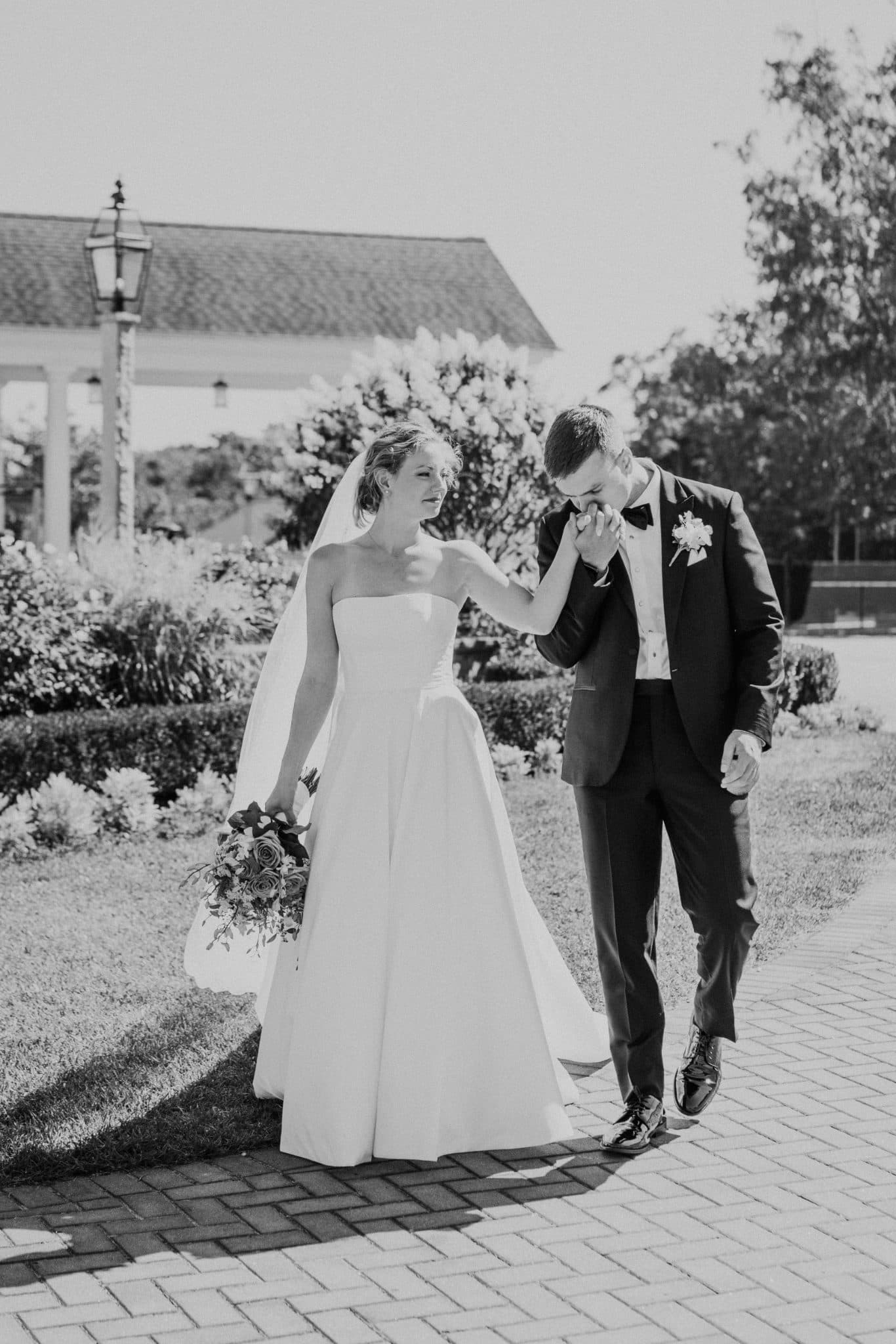 A bride in a strapless gown and veil walks alongside a groom in a suit with a boutonniere, holding hands on a brick path with a garden and building in the background.